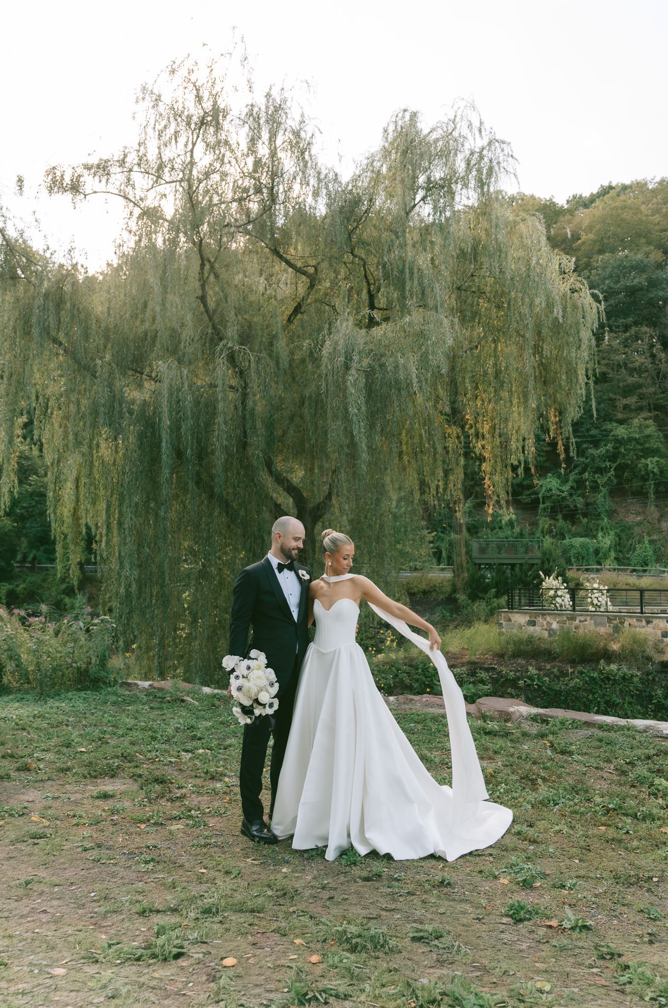 A couple in wedding attire poses together under weeping willow trees at an outdoor venue during their special day.