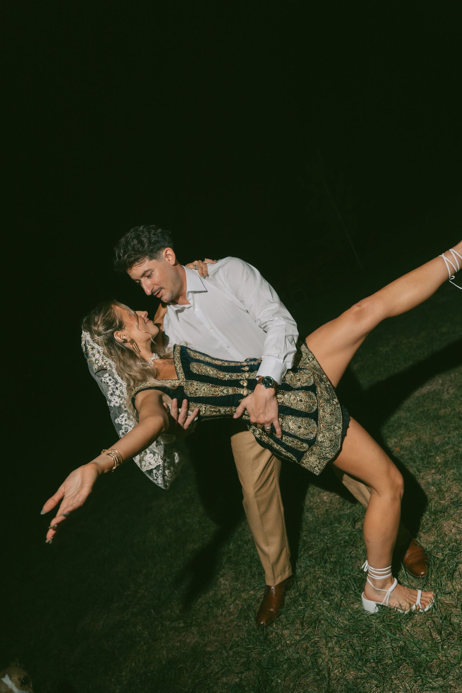Two dancers perform a dramatic dip move on dark grass in traditional Bavarian attire.