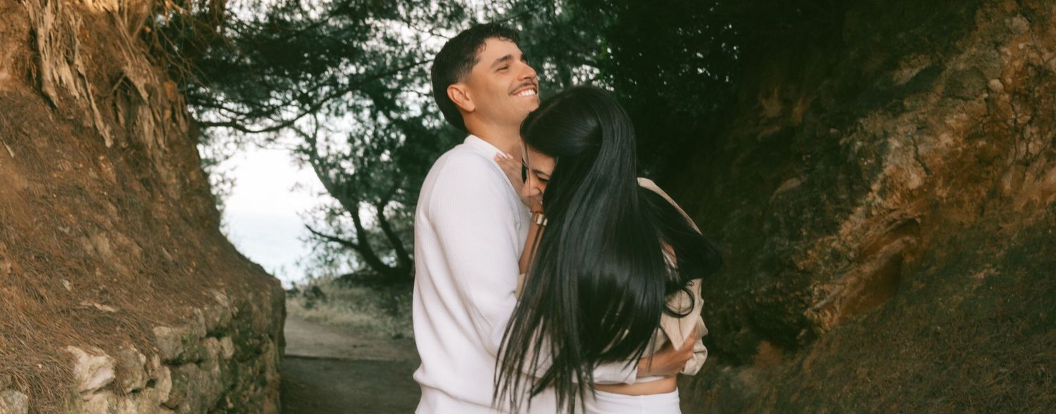 Couple embracing romantically outdoors near rocky cliffs and trees in a tender moment.