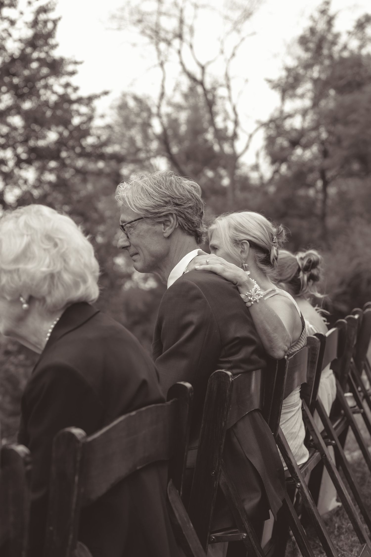 Black and white photograph of people seated at an outdoor gathering in a wooded setting.