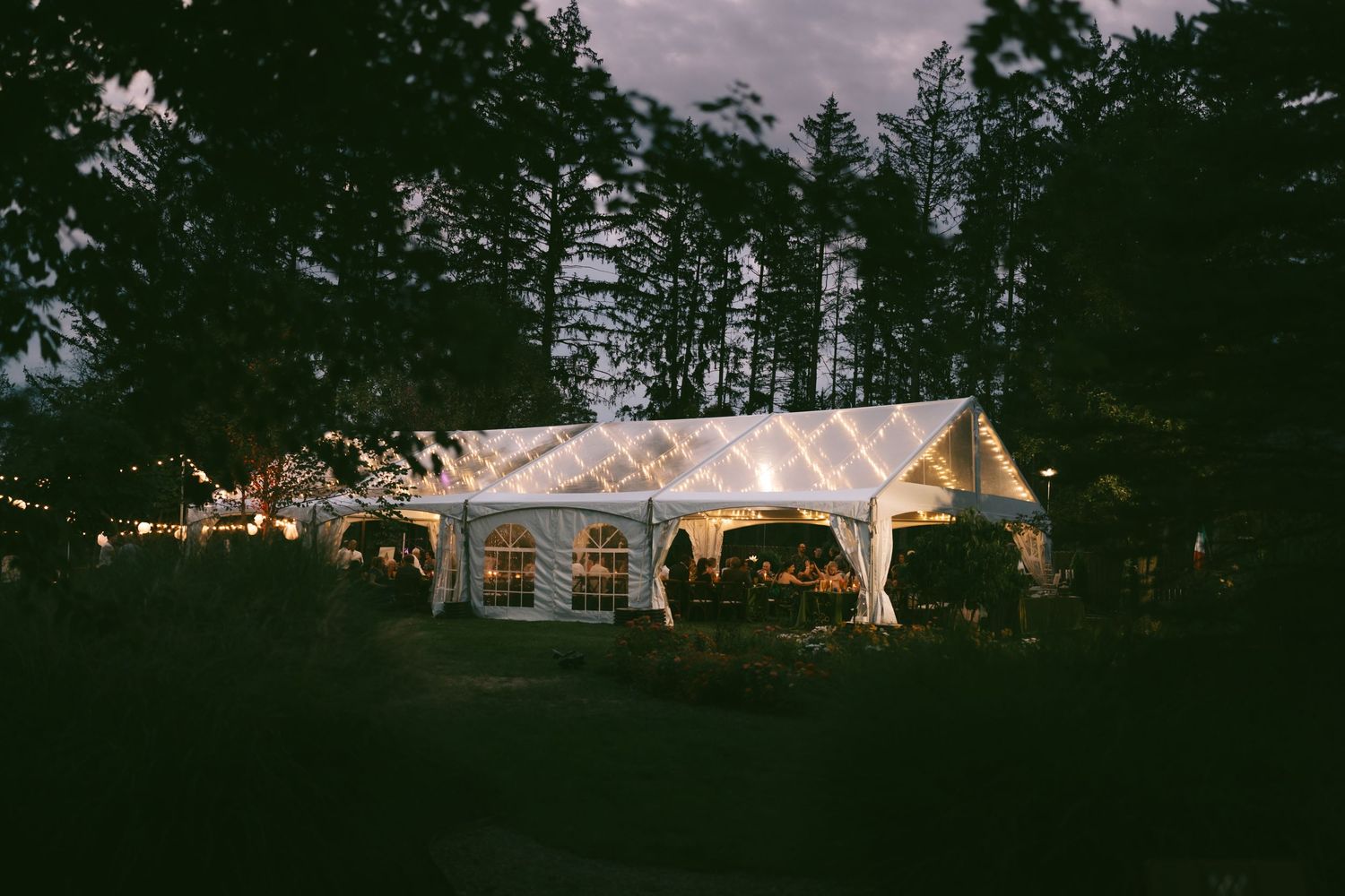 White wedding tent illuminated at dusk with purple sky and trees in the background.