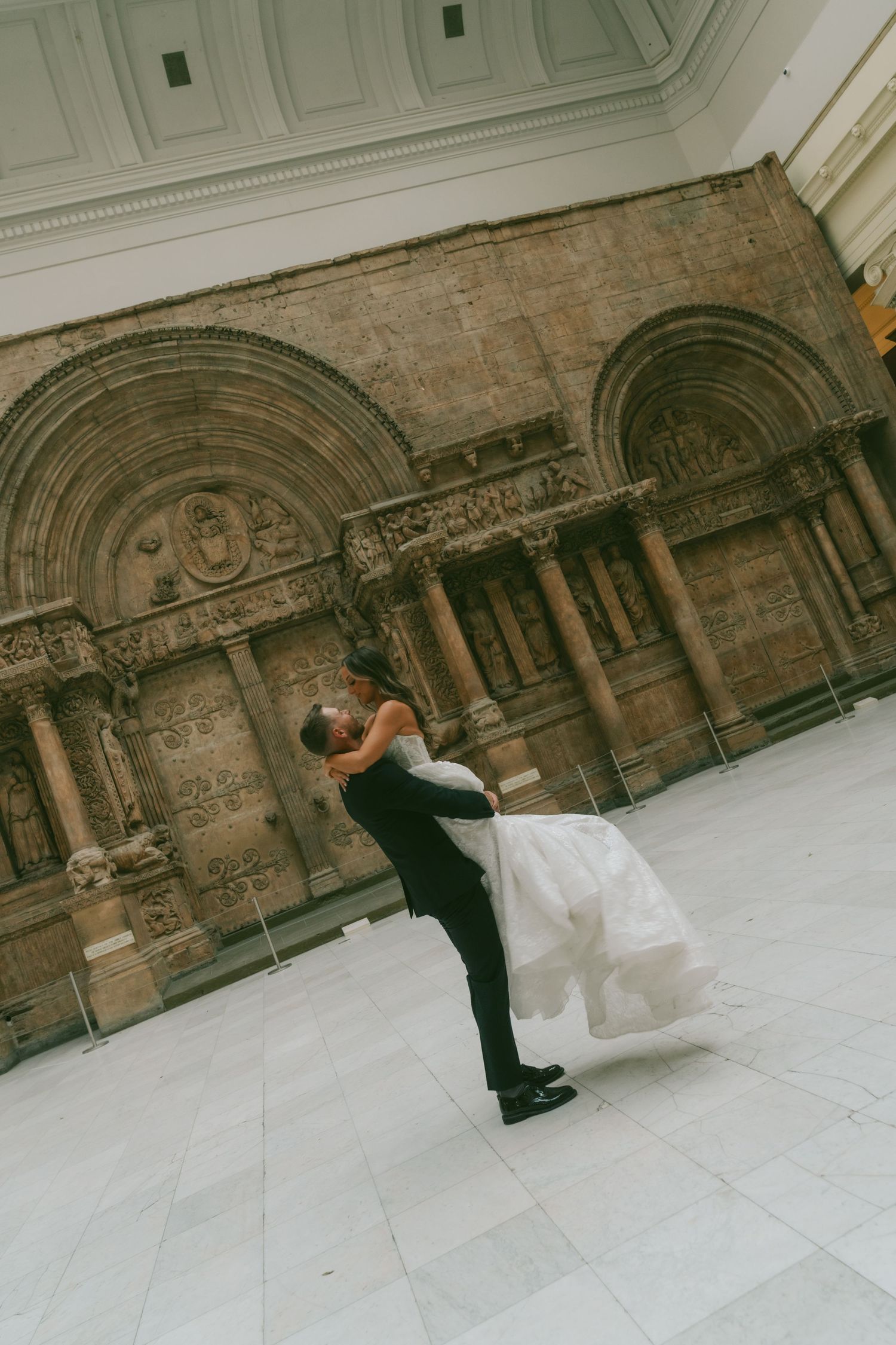 A couple in wedding attire performs a romantic dip pose in front of an ornate stone building facade.