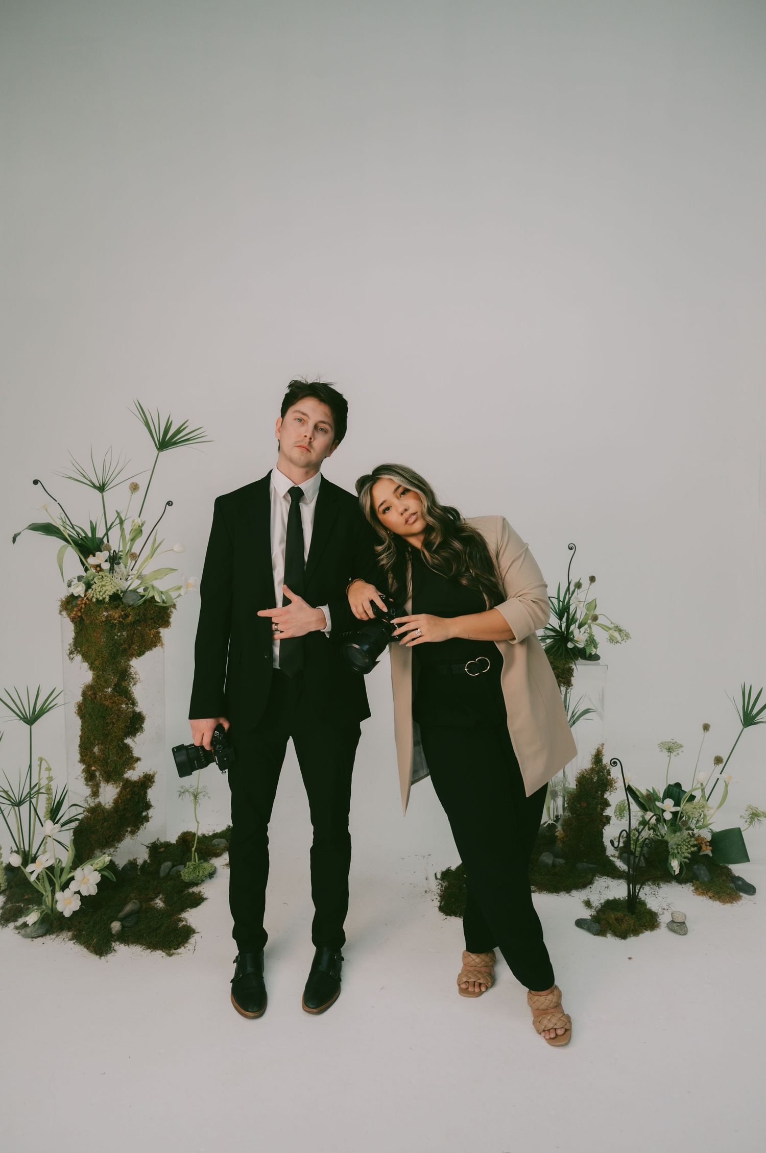 Creative studio photoshoot featuring couple posing playfully with decorative plants against white background.