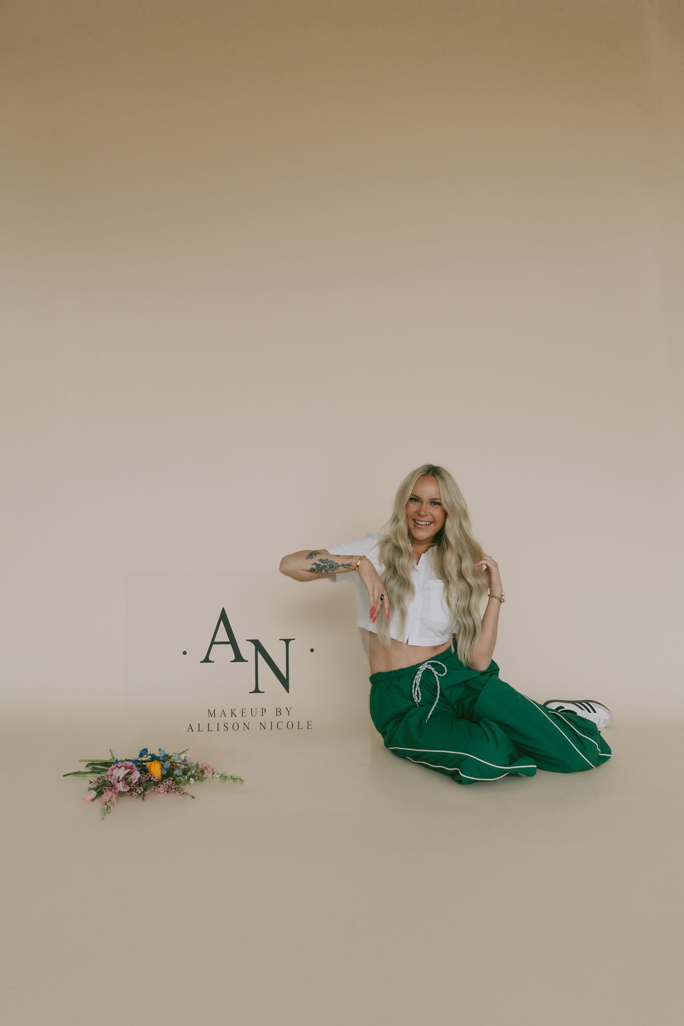 A person in a white crop top and green flowy pants sits on the floor next to decorative flowers and a logo.