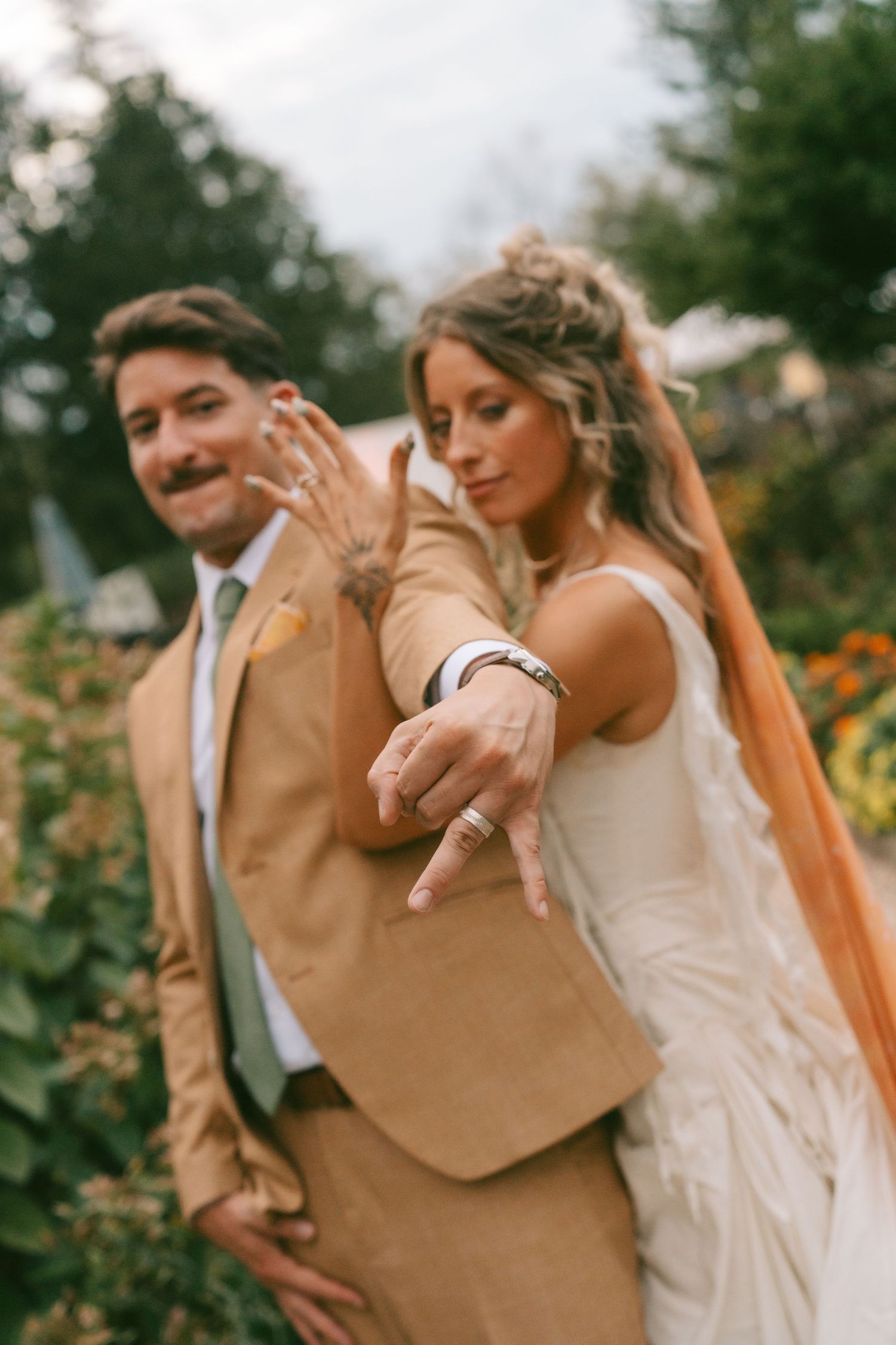 A couple in wedding attire showing off their rings in a garden setting at sunset.