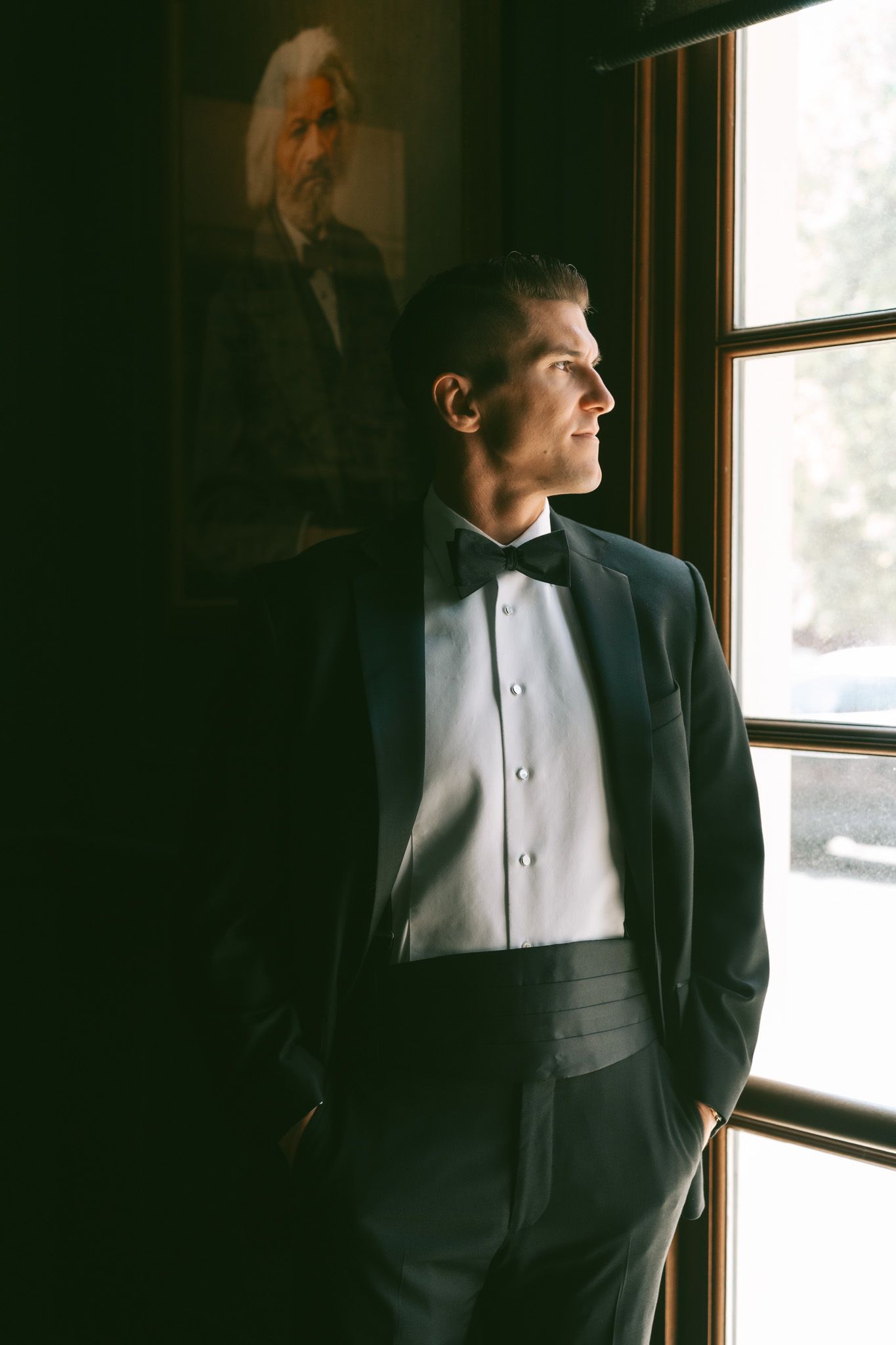 A person in a formal black tuxedo and bowtie stands contemplatively by a window with dramatic lighting.