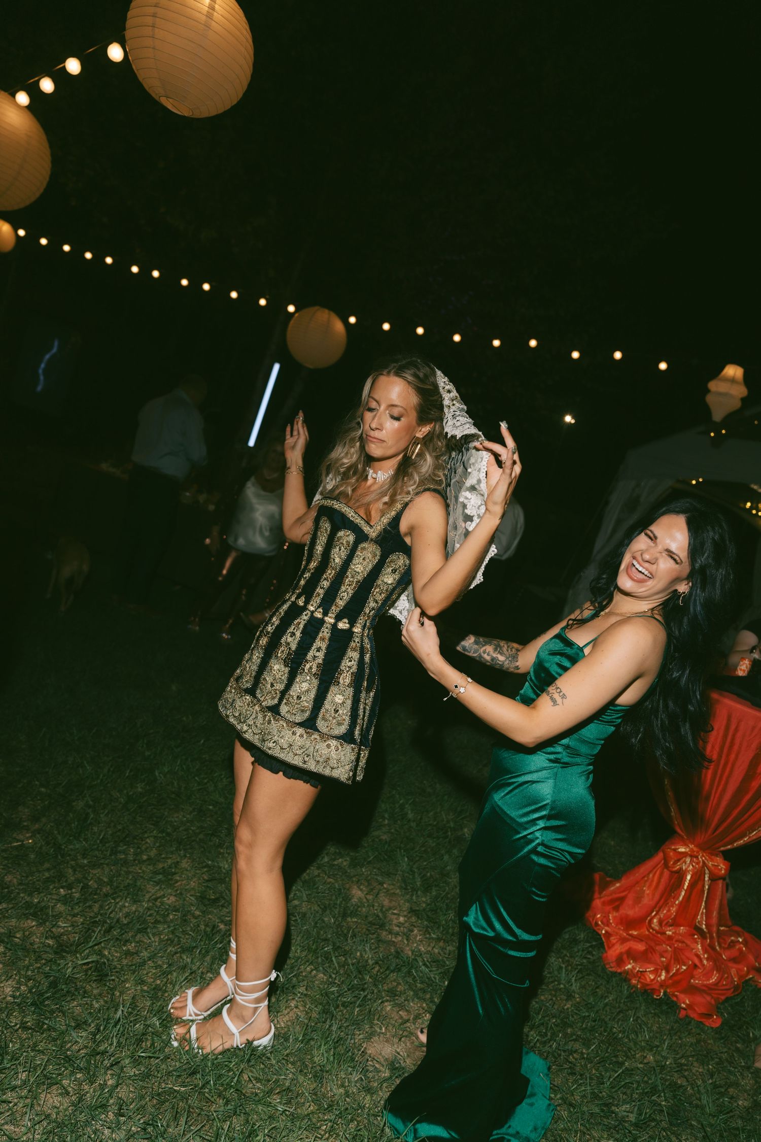 Two partygoers dance energetically at a nighttime outdoor celebration with paper lanterns and string lights overhead.
