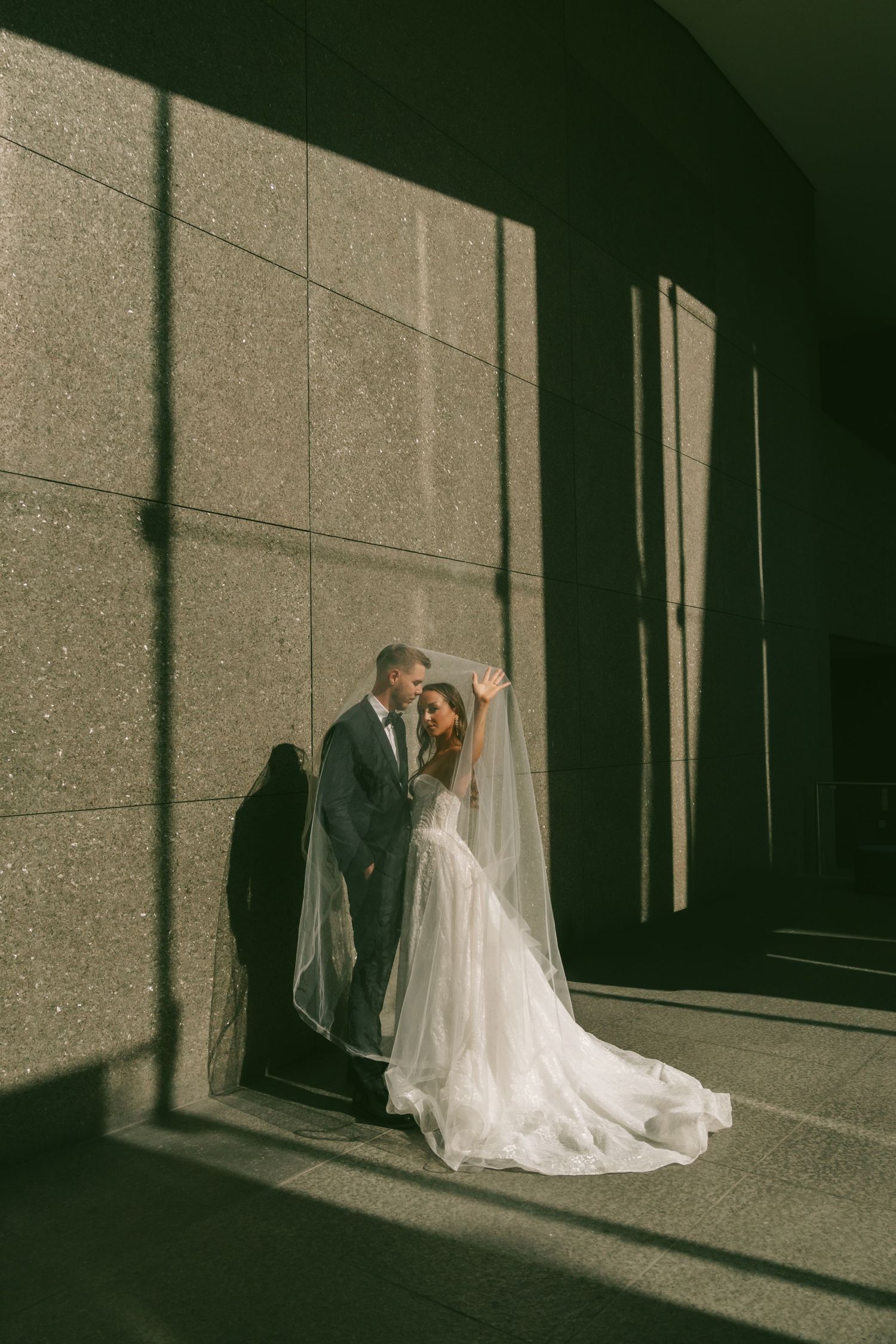 Dramatic shadows cast on stone walls as newlyweds pose in formal wedding attire.