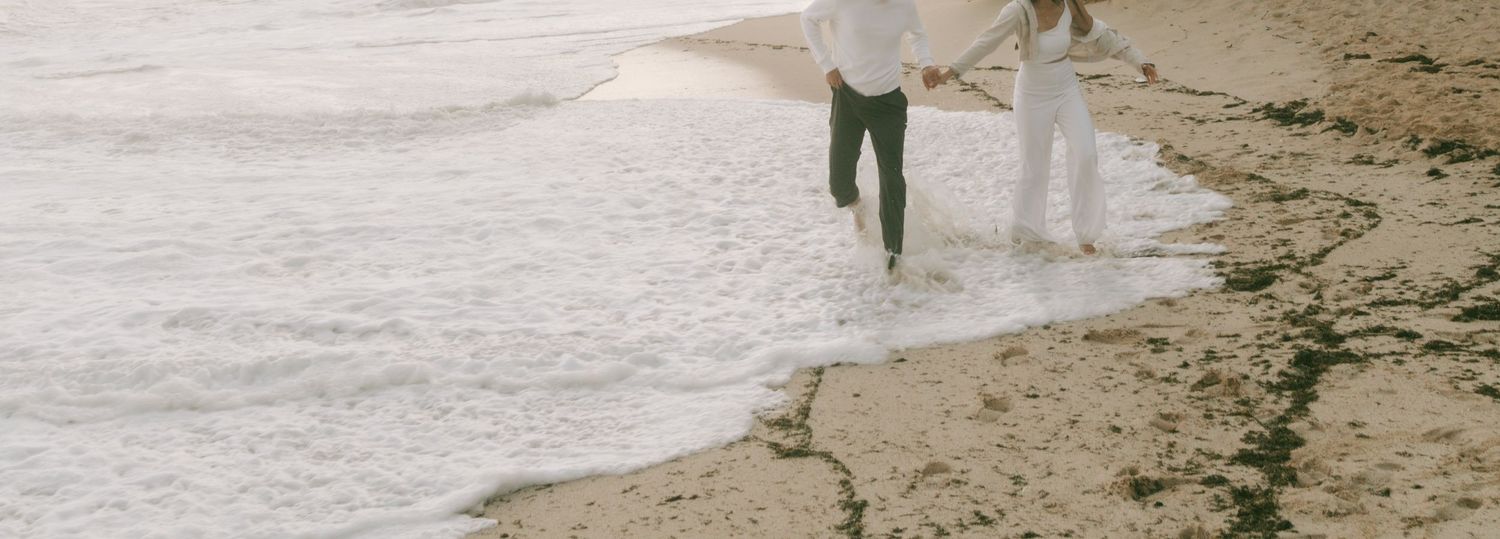 Couple walking hand in hand along a sandy beach shore with gentle waves, wearing casual white and green outfits.