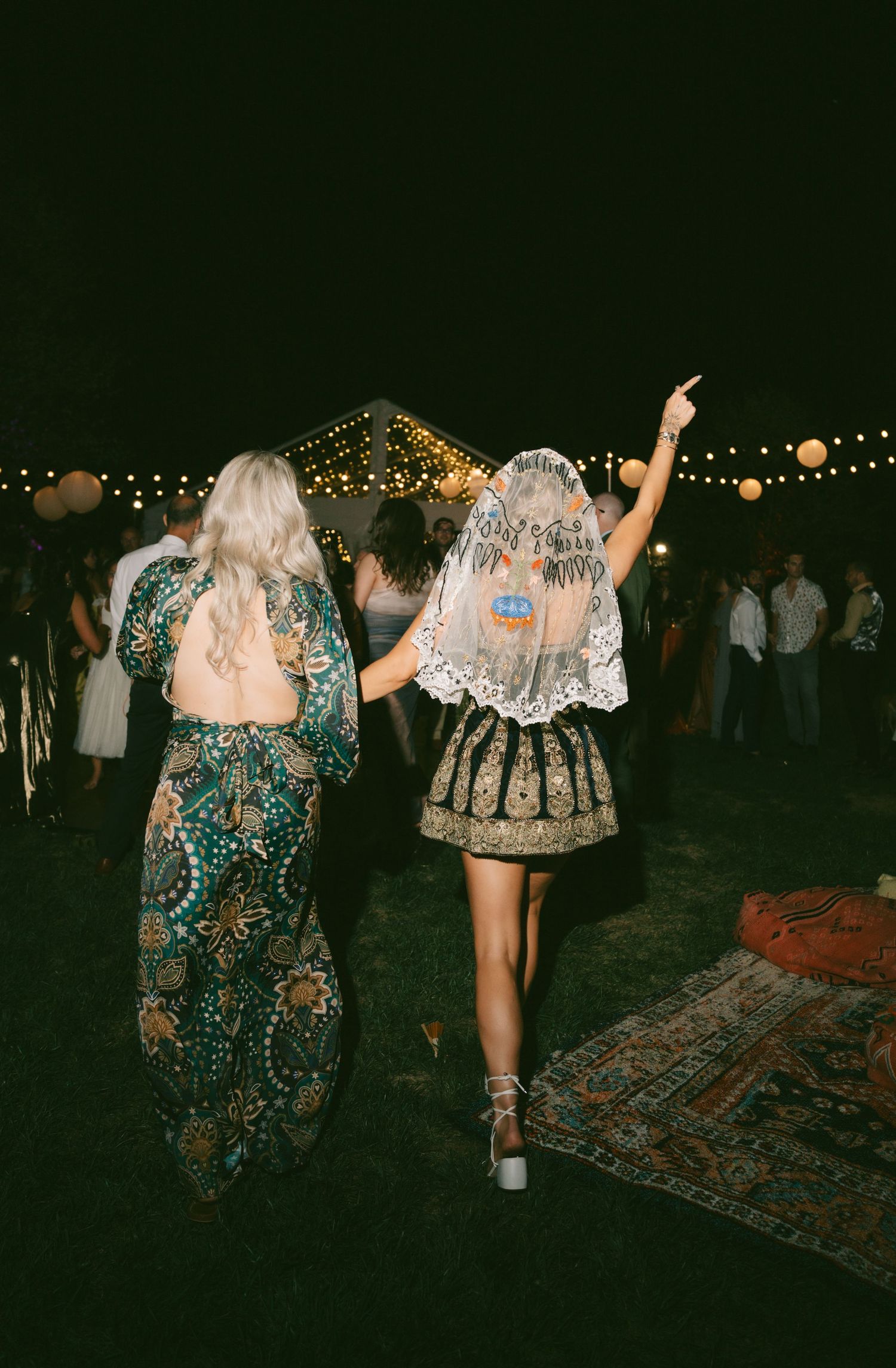 Partygoers in sparkly dresses dancing under string lights at night.