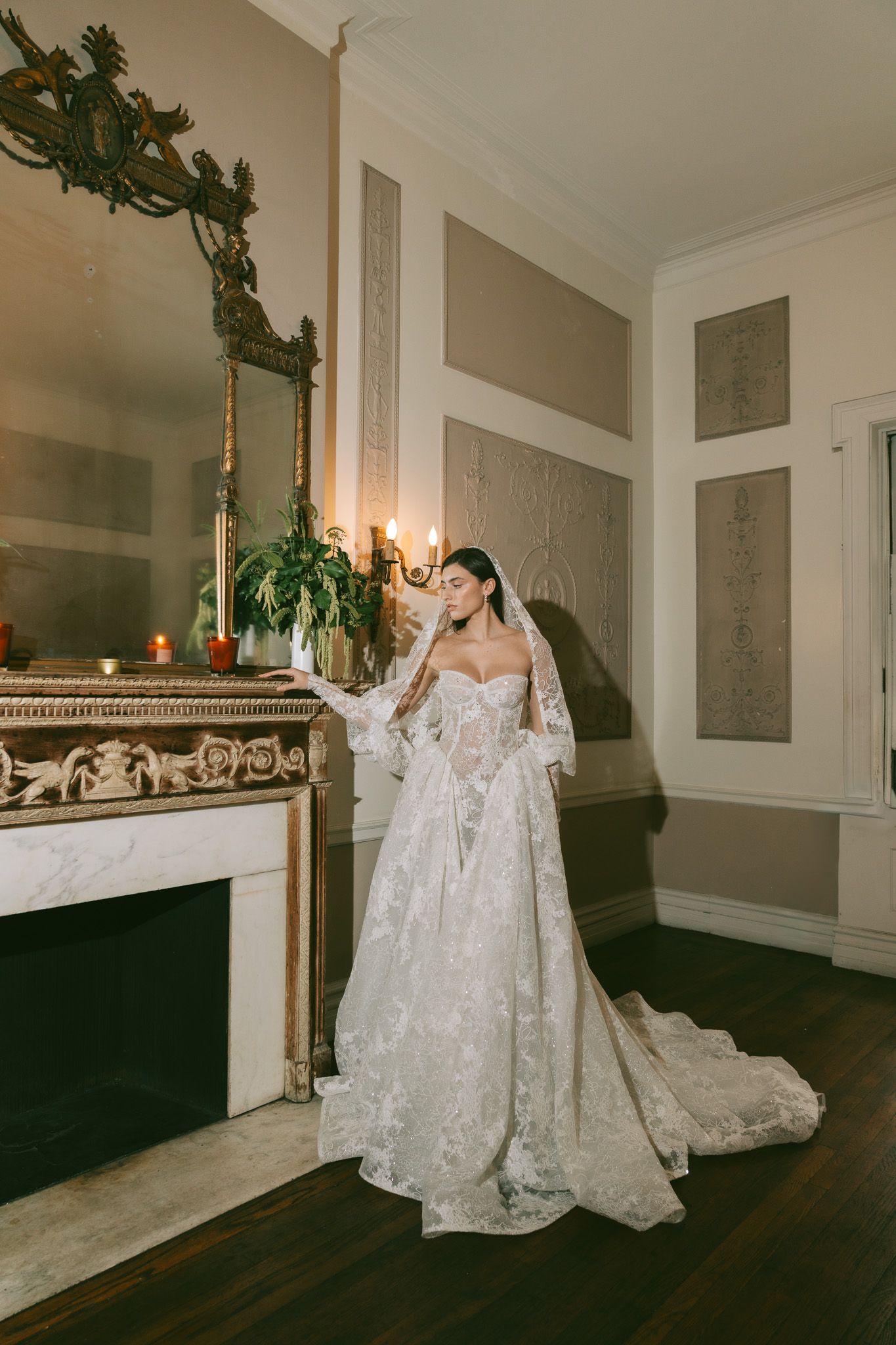 Elegant white lace wedding gown with off-shoulder design and flowing train photographed in vintage room with ornate fireplace.