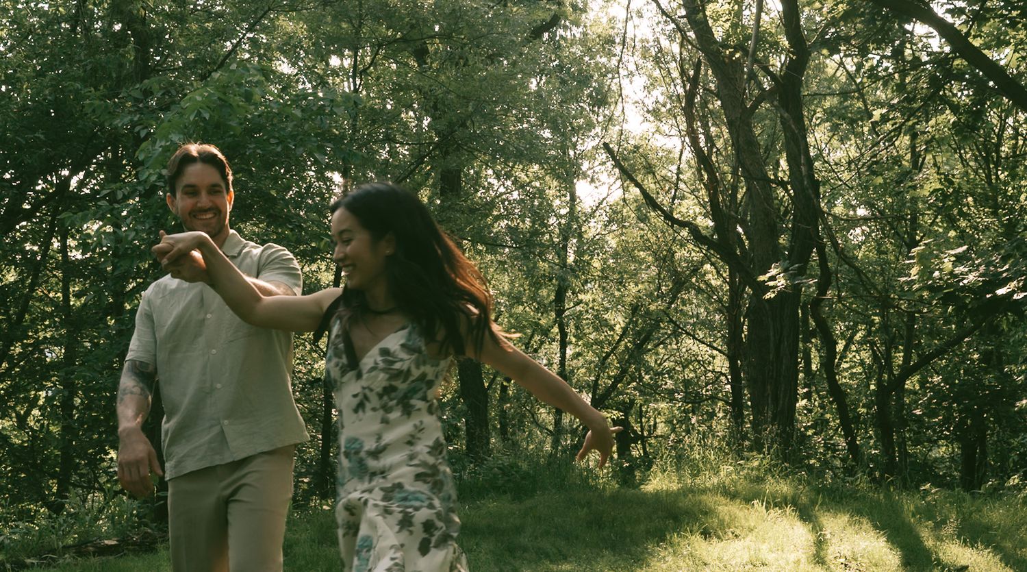 A couple dances joyfully together in a sunlit forest setting.