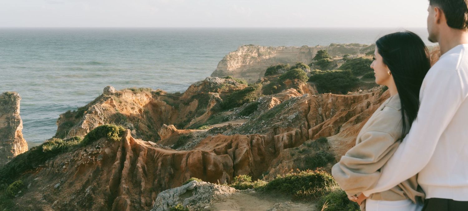 Couple standing together overlooking dramatic coastal cliffs and ocean at golden hour sunset.