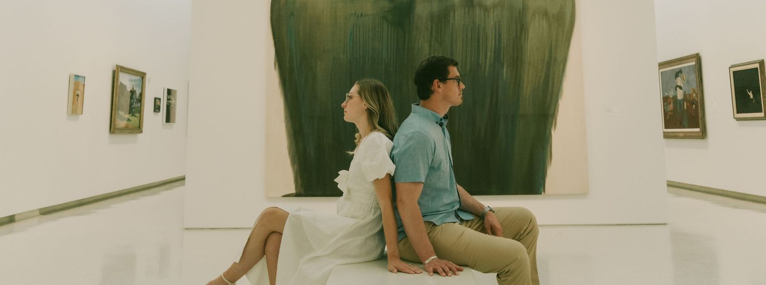 Two people sit back-to-back on a wooden bench in an art gallery with framed pieces on white walls.