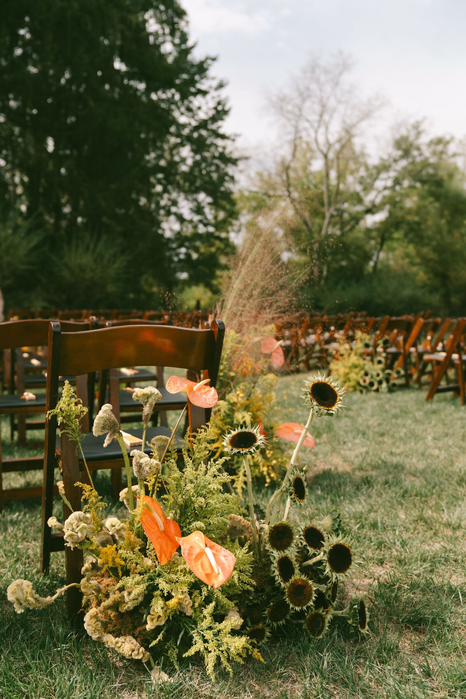 Rustic outdoor wedding ceremony setup with wooden chairs and sunflower floral arrangements in a garden setting.