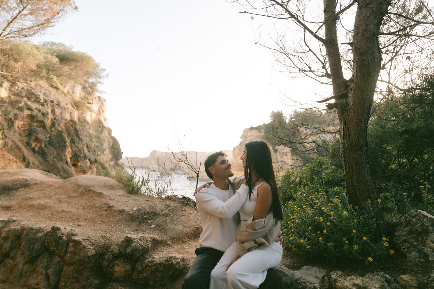Couple sitting on rocky coastal cliffside surrounded by lush greenery, sharing a tender romantic moment in warm golden light.
