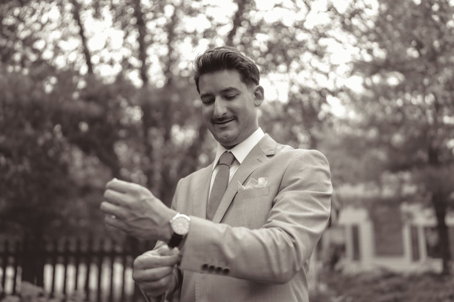 A person in a light colored suit adjusts their watch while standing outdoors in a black and white photograph.