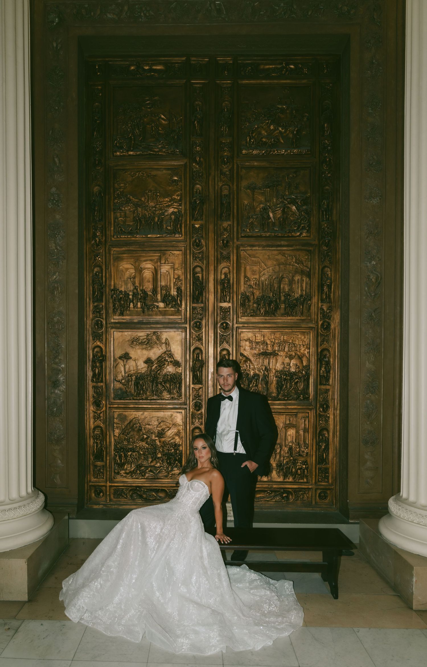 Elegant bride in flowing white gown and groom in tuxedo pose in front of ornate carved wooden panels in grand hall.