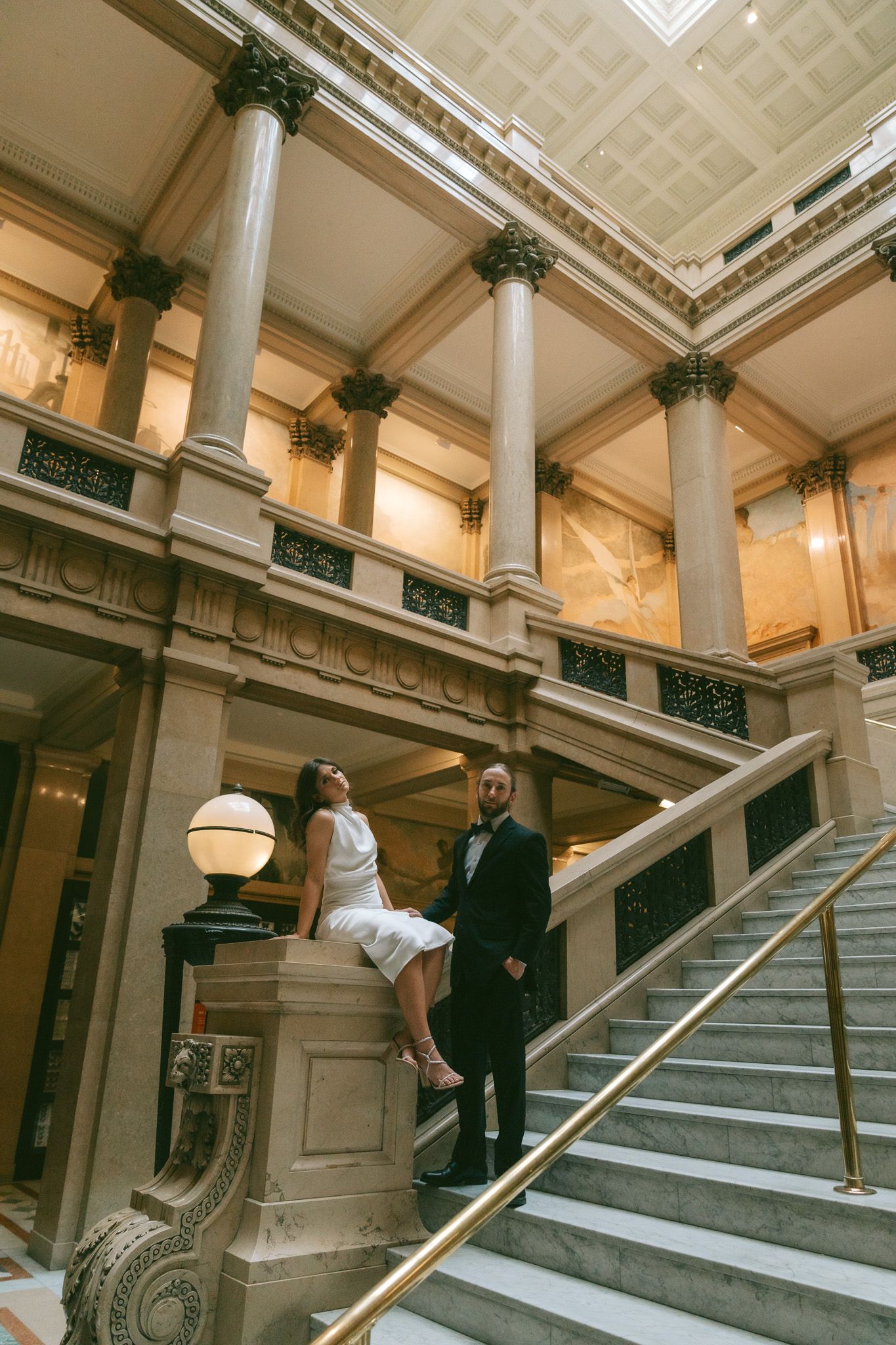 A couple poses on elegant marble stairs inside a grand historic building with ornate columns and architectural details in carnegie museum