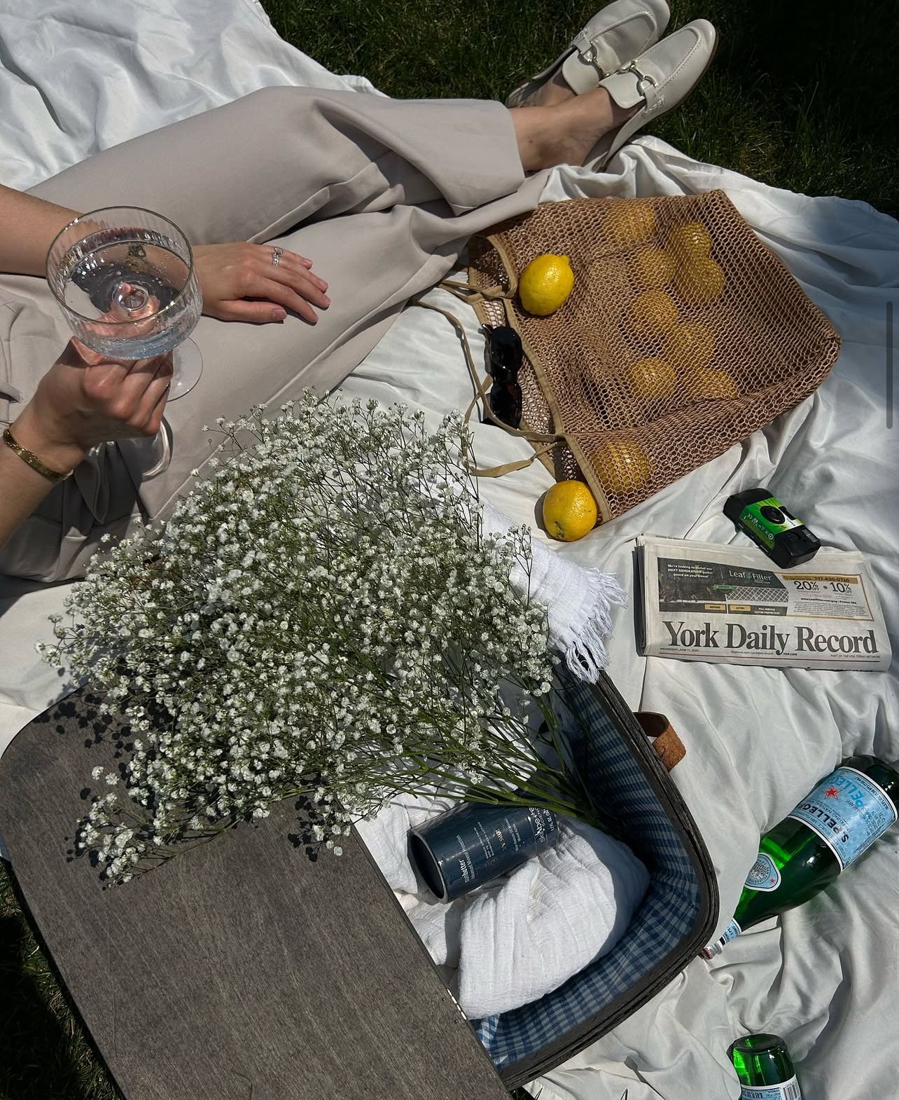 Picnic setup with baby's breath flowers, newspaper, and drinks on white blanket.