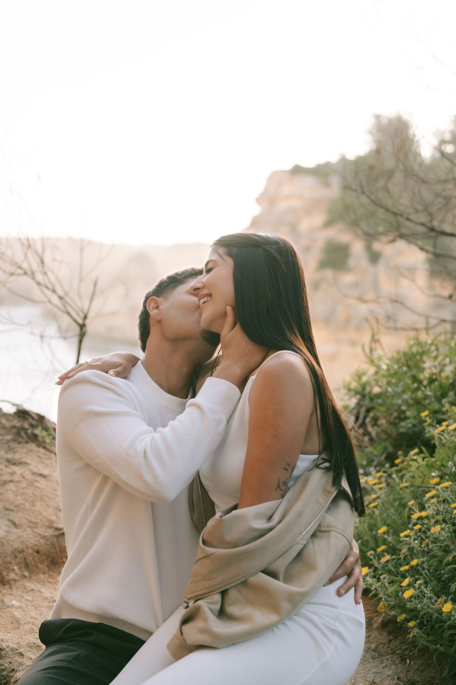 Couple sharing a romantic kiss outdoors at golden hour with rocky cliffs and greenery in the background.