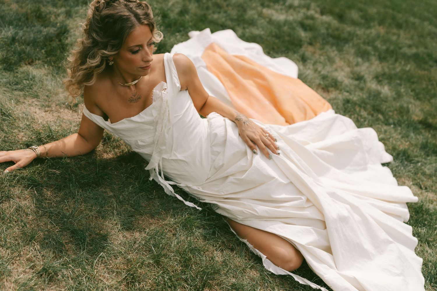 A person in a flowing white dress lies gracefully on grass during an artistic outdoor photoshoot.