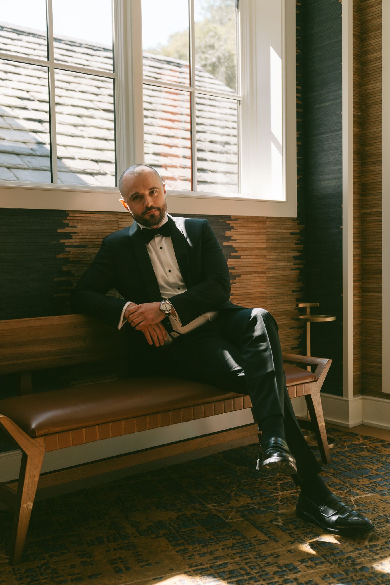 A person in a black tuxedo sits elegantly on a brown leather bench near a window in warm lighting.