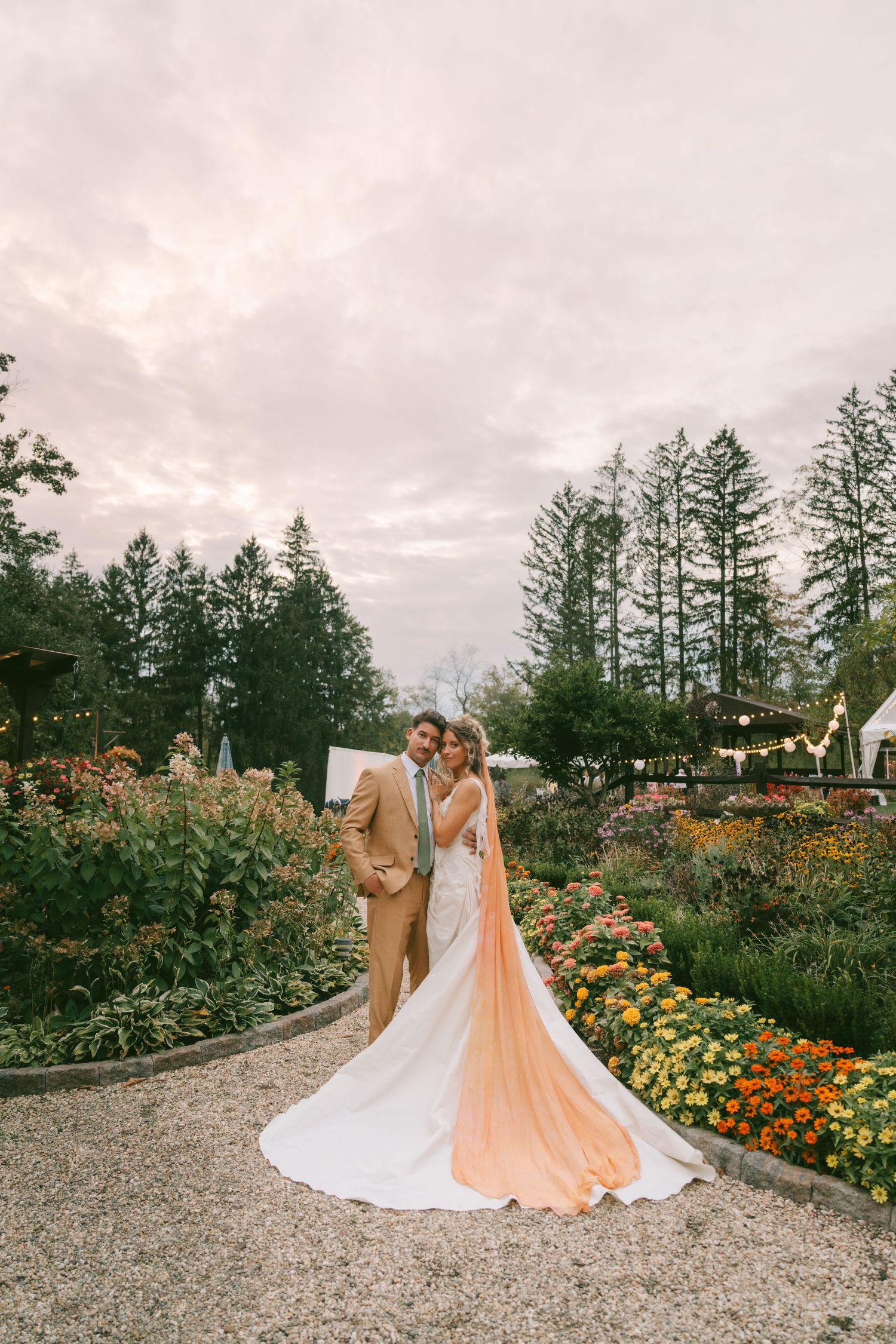 Wedding couple poses in a garden filled with colorful flowers and evergreen trees.
