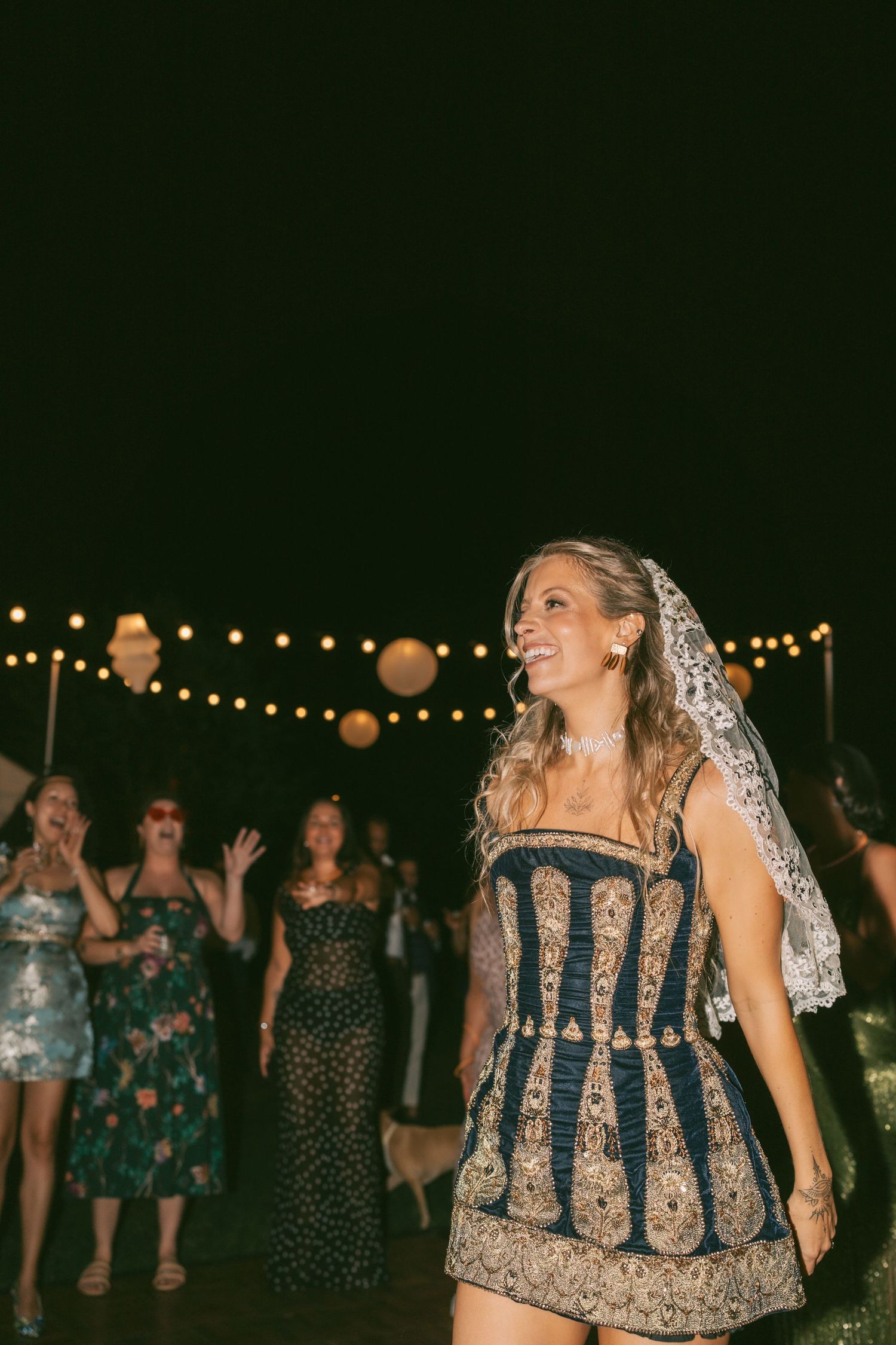 People dancing and celebrating at a nighttime wedding reception under string lights.