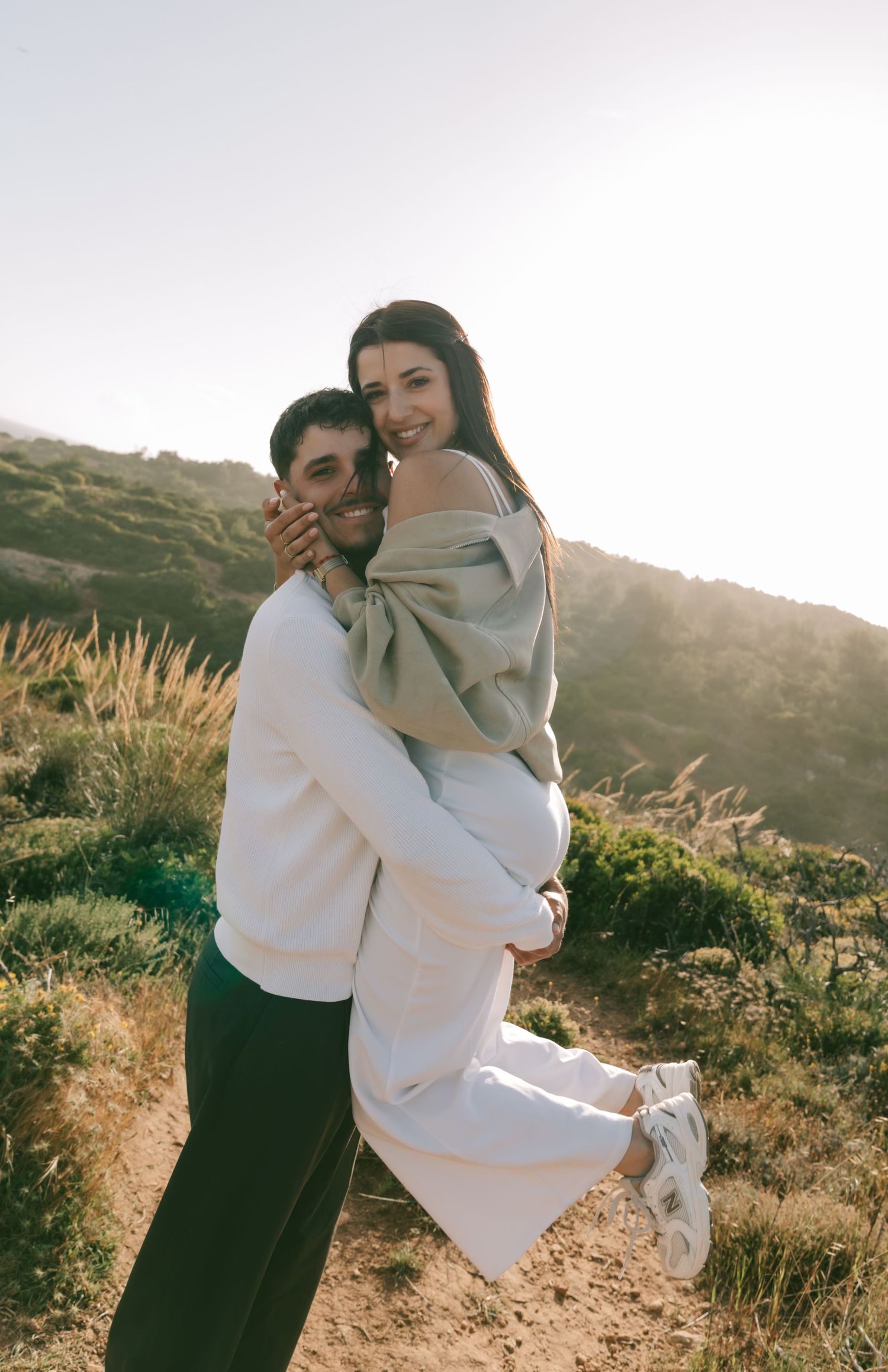 A couple embraces outdoors on a sunny hillside, the man lifting the woman as they smile warmly at the camera.