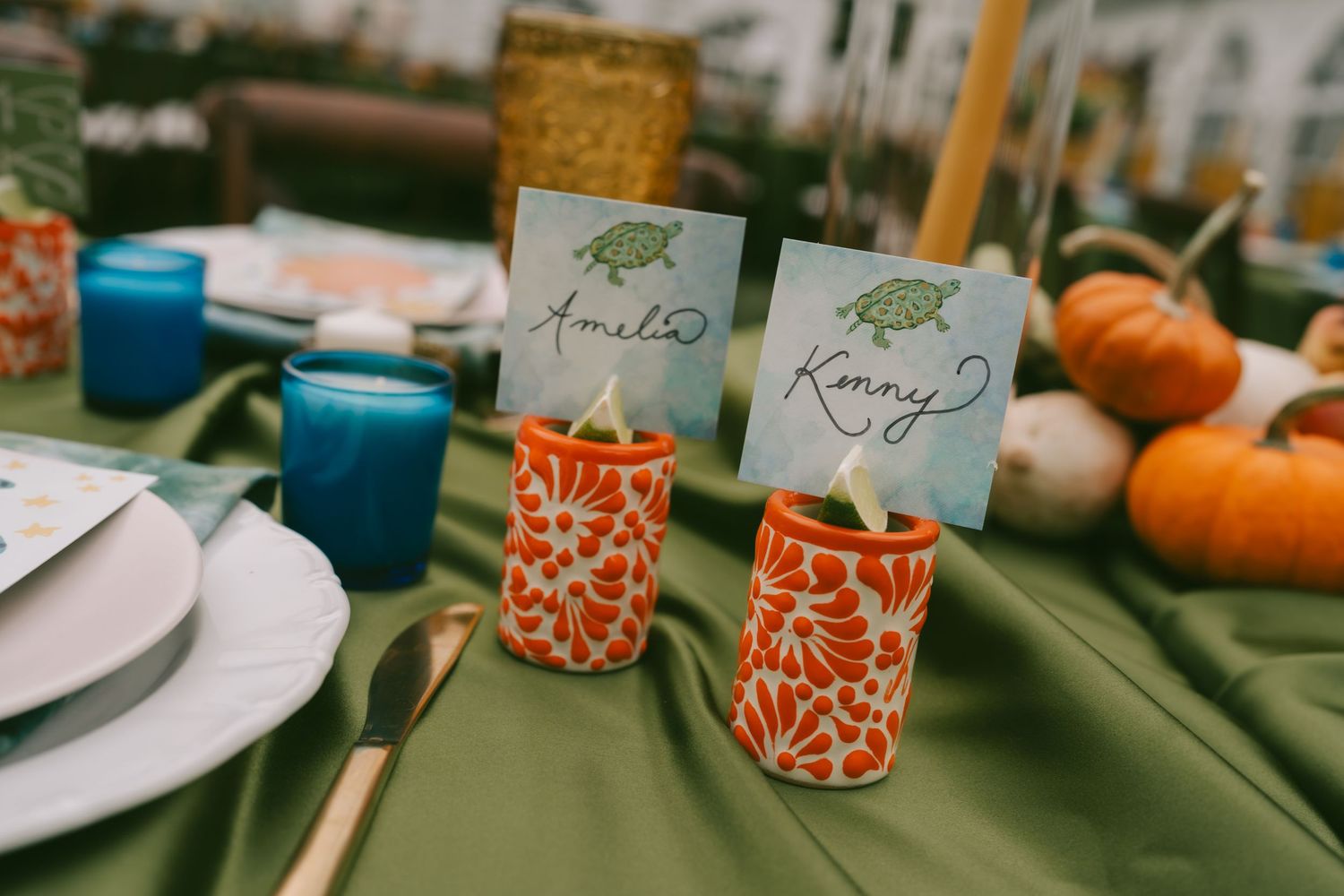 Elegant table setting with orange candle holders, teal candles, and autumn decor arranged on a green tablecloth.