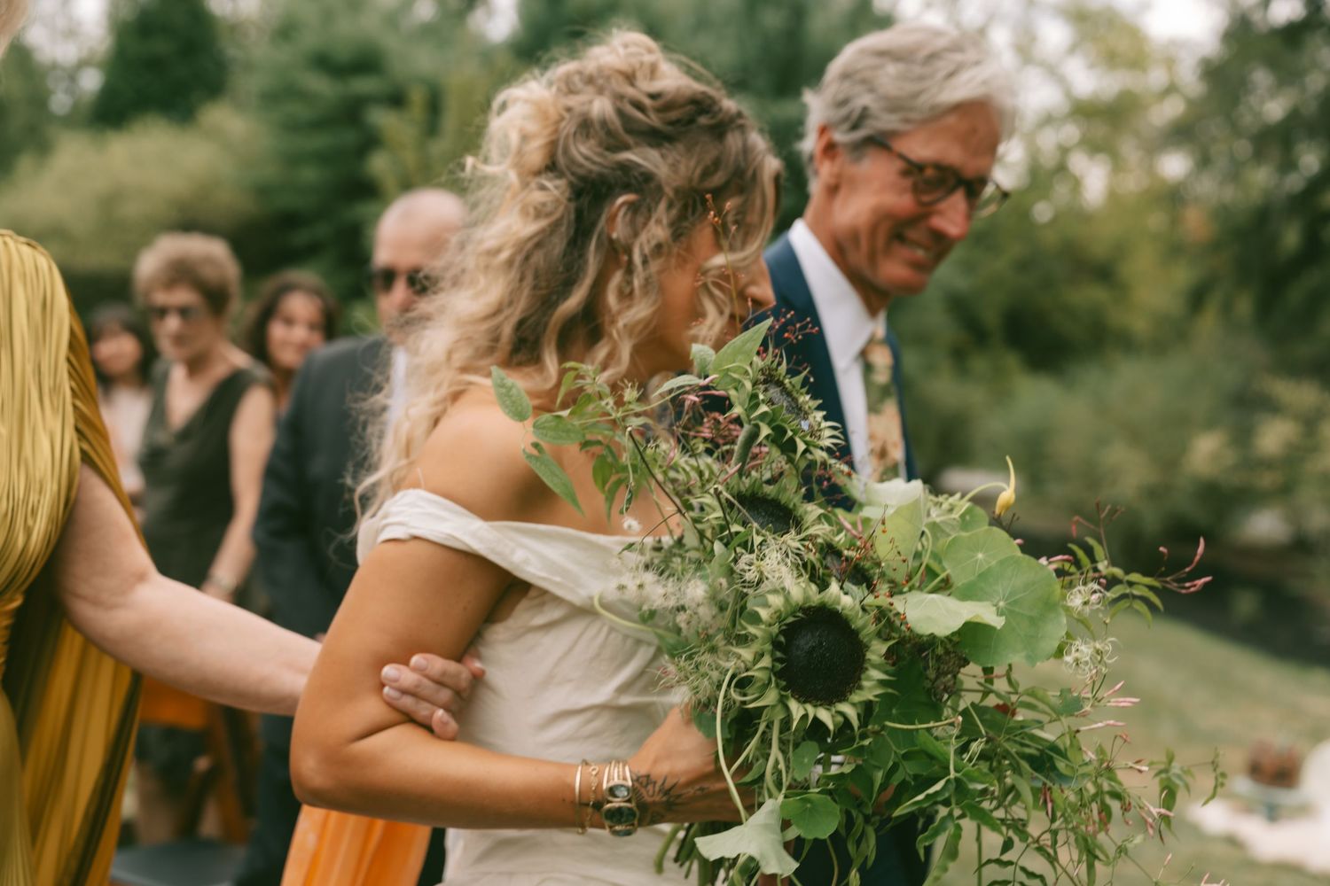 A bride in an off-shoulder dress holds a wild green bouquet during an outdoor wedding ceremony.