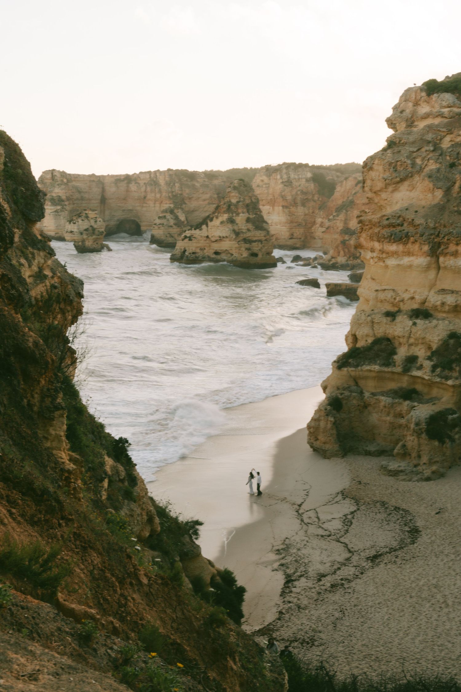 Two people walking on a secluded sandy beach surrounded by towering golden limestone cliffs in the Algarve, Portugal.