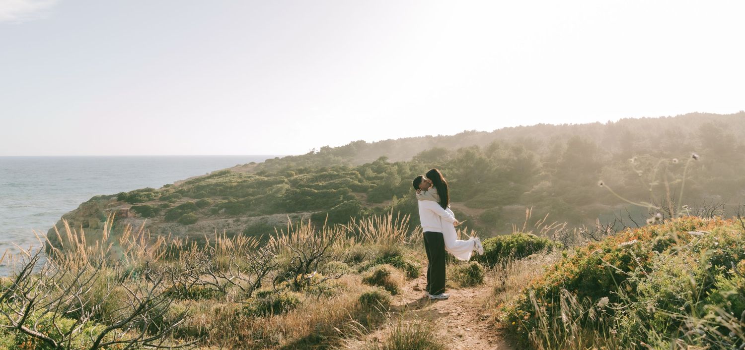 Couple embracing on a scenic coastal cliff path surrounded by lush greenery with ocean views at golden hour.