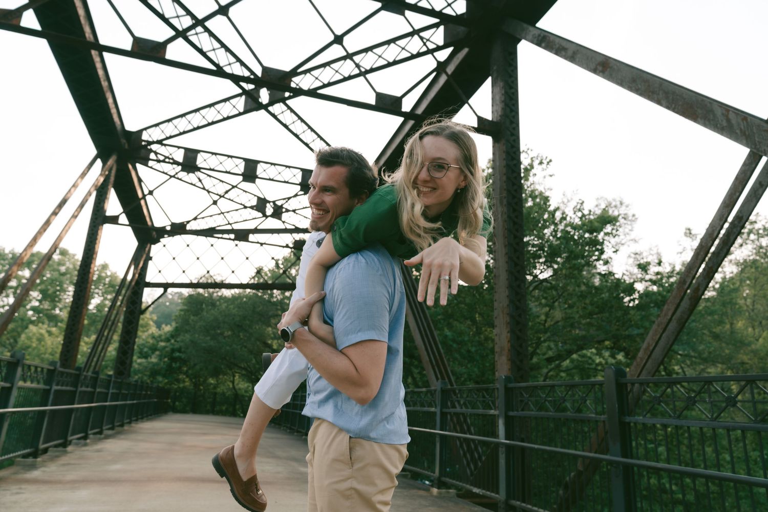 Two people playfully pose back-to-back on a metal bridge structure surrounded by greenery.