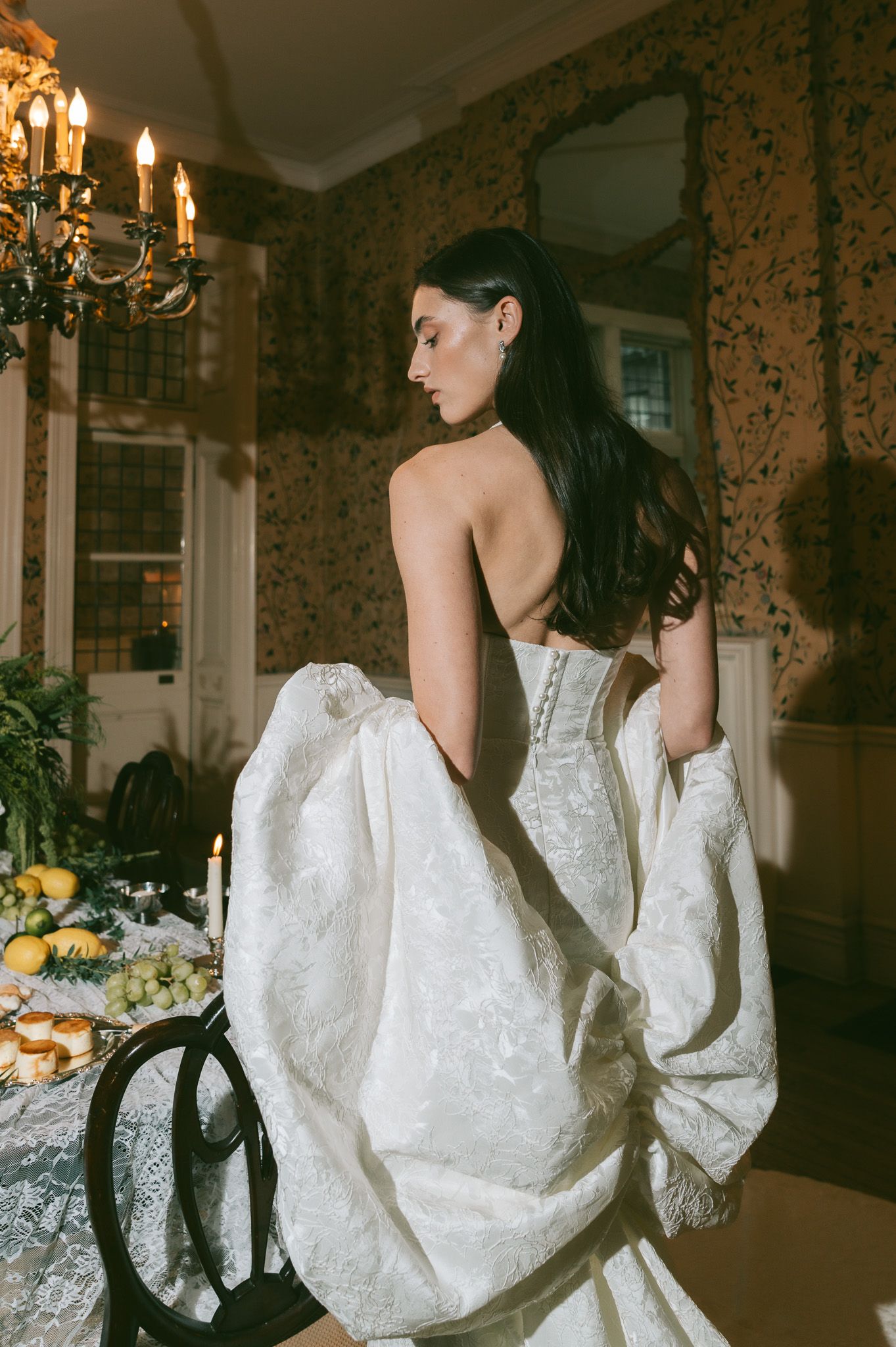 A bride in a white corset wedding dress poses in an elegant room with vintage wallpaper and chandelier lighting.