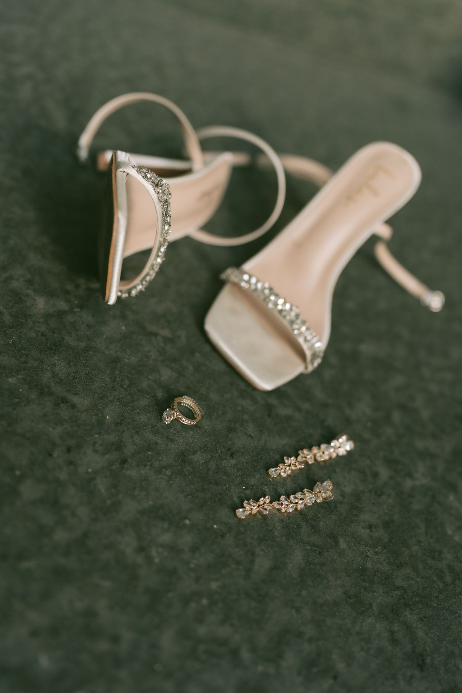 Wedding accessories including cream high heels, jewelry and rings arranged on dark velvet surface.