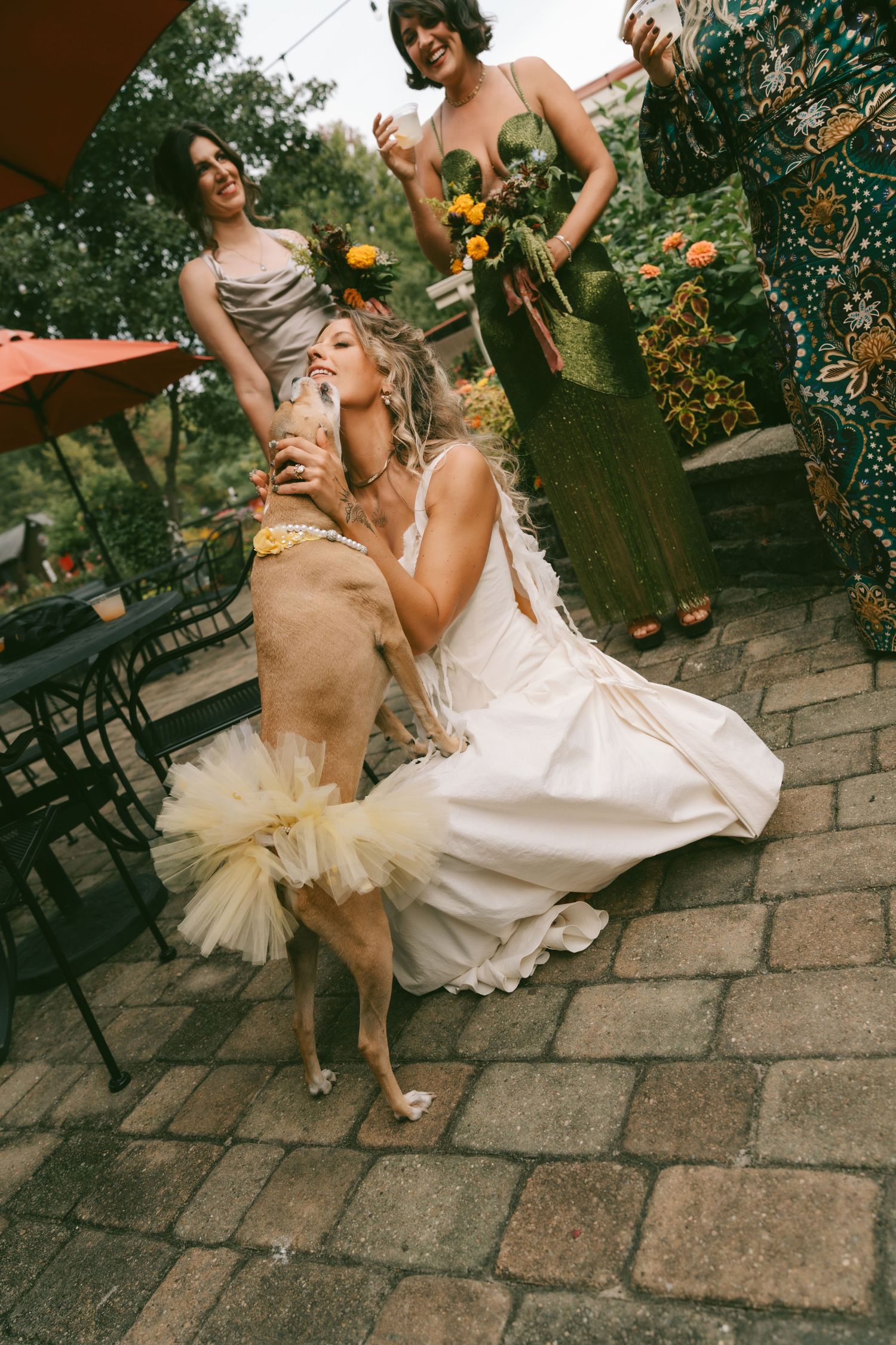 Wedding party members in dresses laugh and dance together on stone pavement during outdoor celebration.