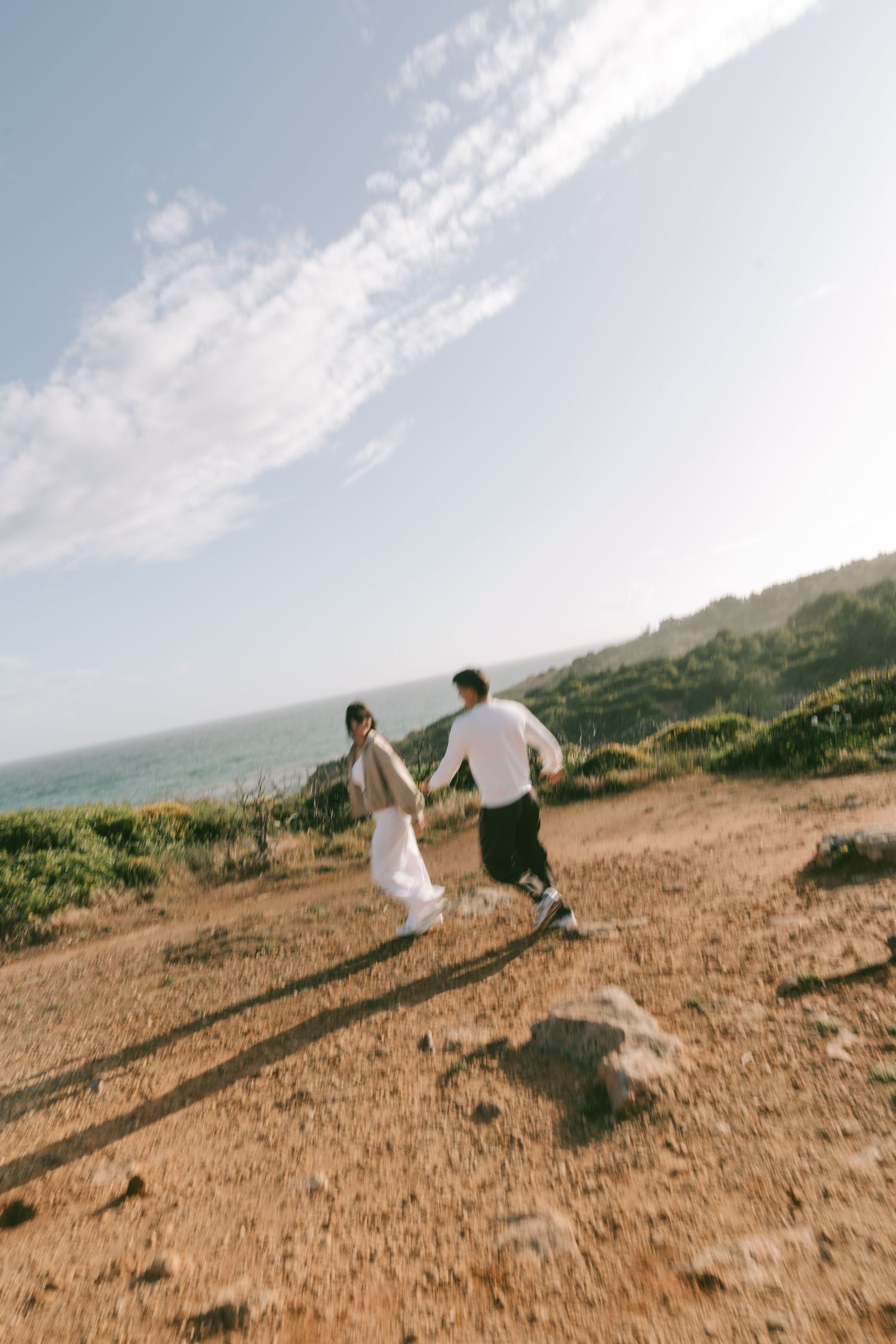 A couple holds hands running on a coastal hillside trail under a bright blue sky with wispy clouds.