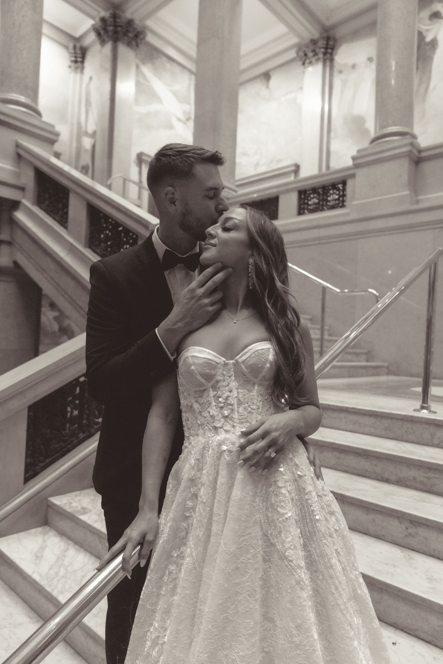 A romantic black and white wedding photo of a couple embracing on an elegant staircase with ornate architecture.