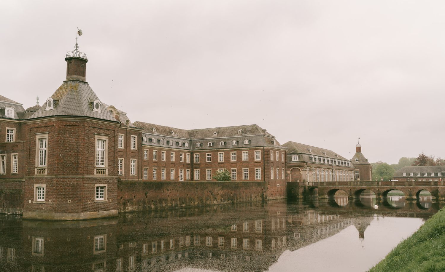Historic brick mansion with octagonal tower reflected in still waters on an overcast day in Germany.