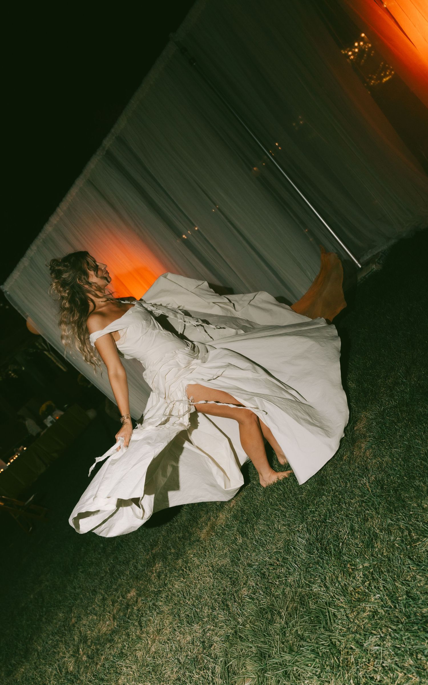 A person in a flowing white dress lies dramatically on grass at night with orange lighting.