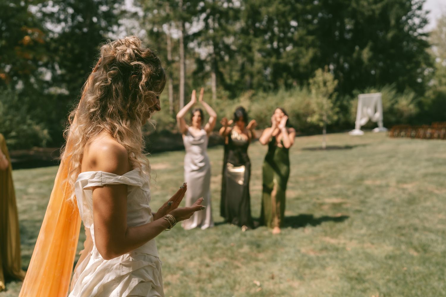 Outdoor wedding ceremony with guests gathered under evergreen trees and orange decor accents.