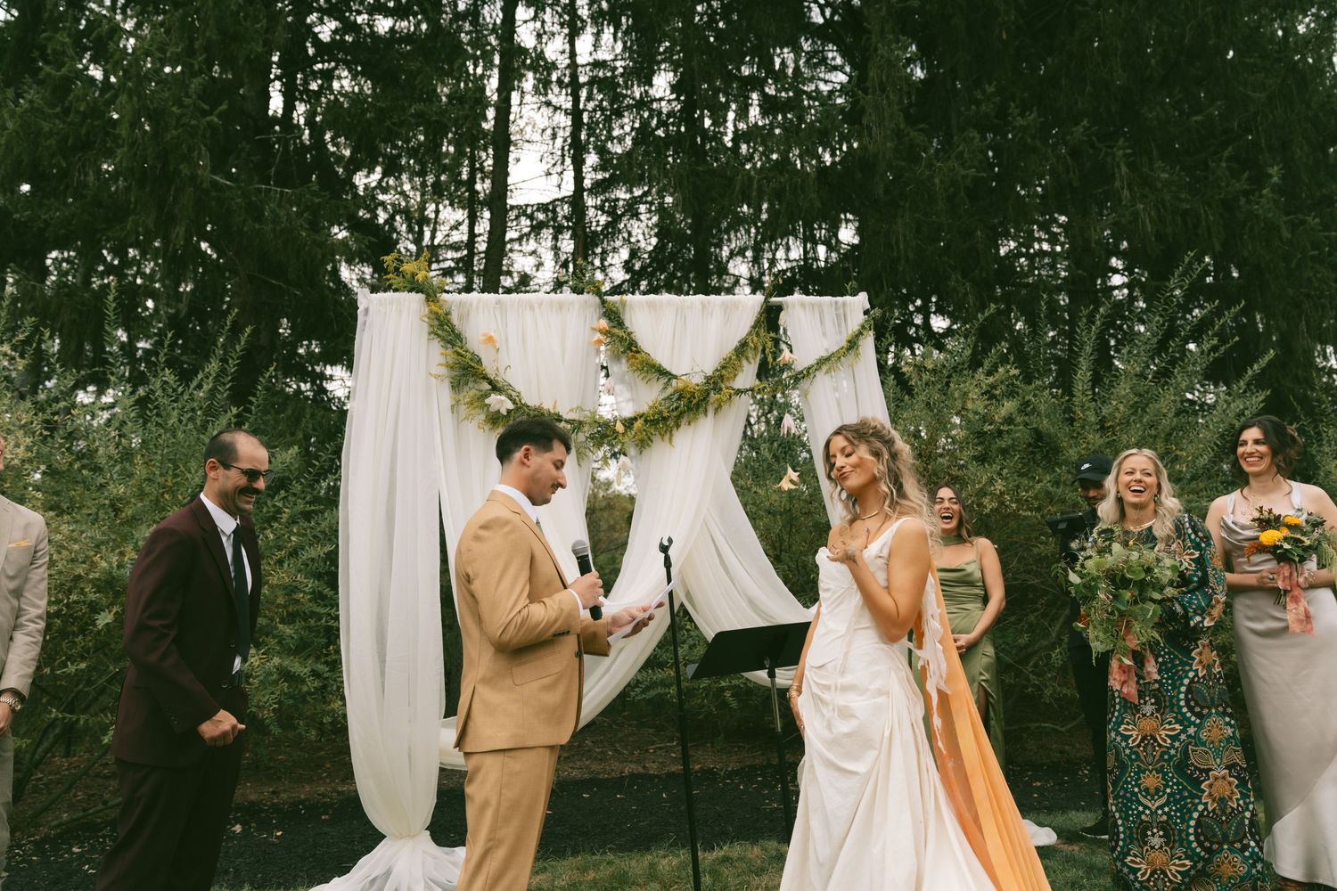 Outdoor wedding ceremony with white draping and greenery in a wooded setting.