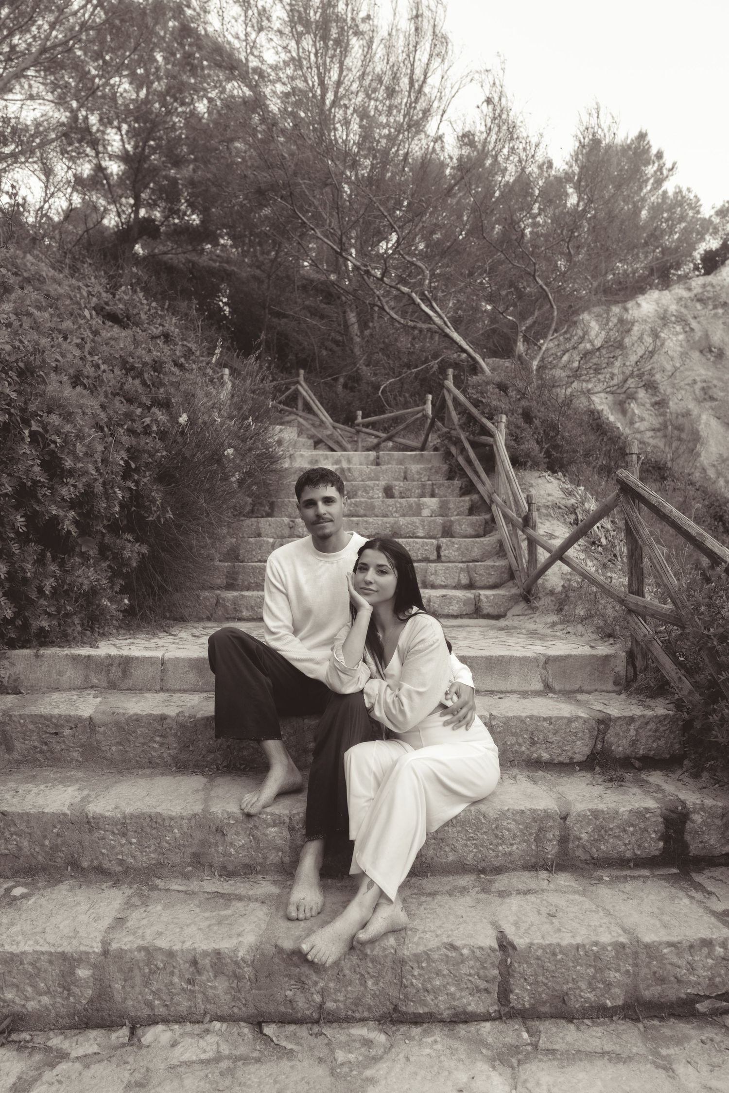 Black and white photo of a couple sitting barefoot on stone steps surrounded by trees and rustic wooden railings.