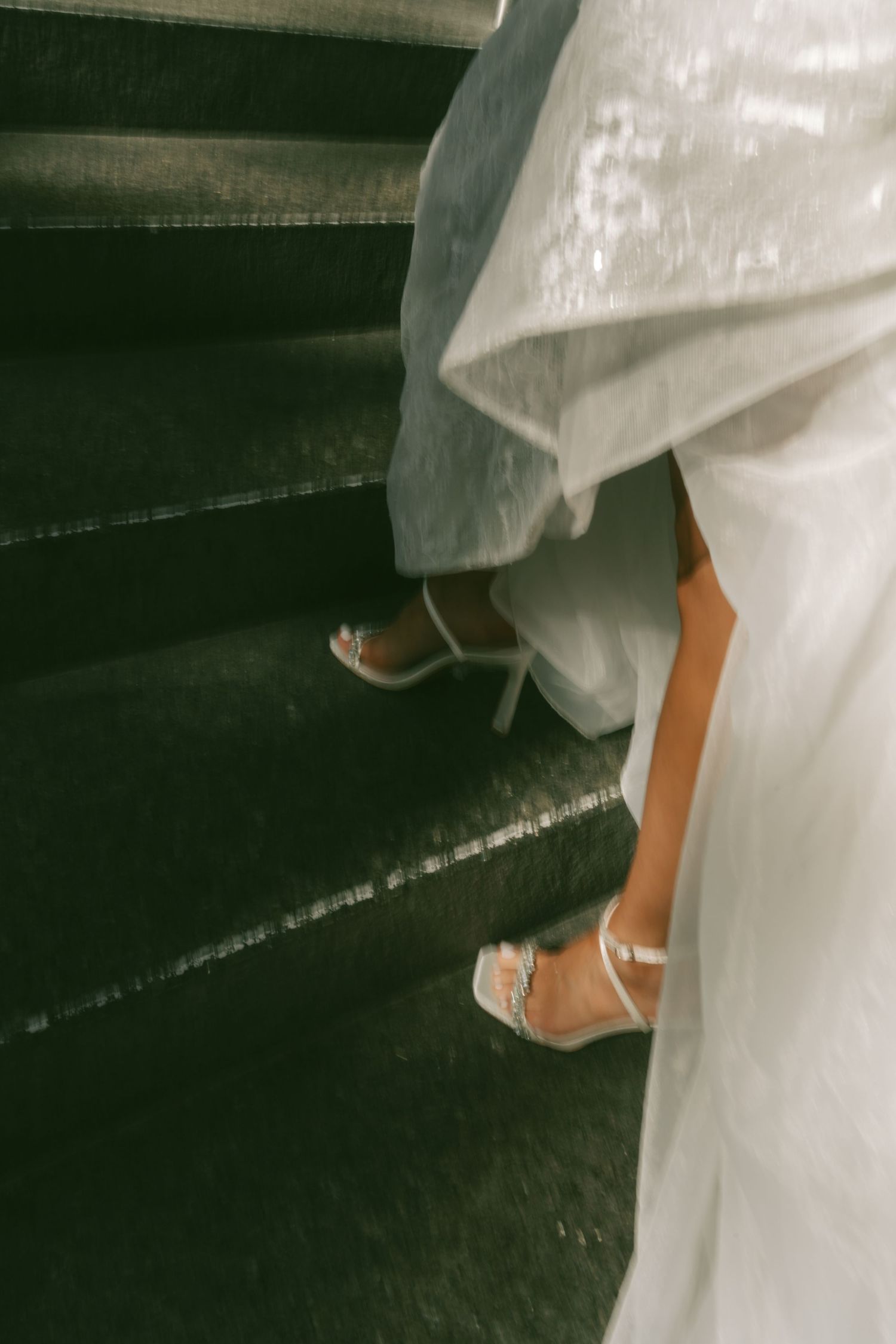 Close-up of bridal shoes and flowing white wedding dress hem against a dark floor.