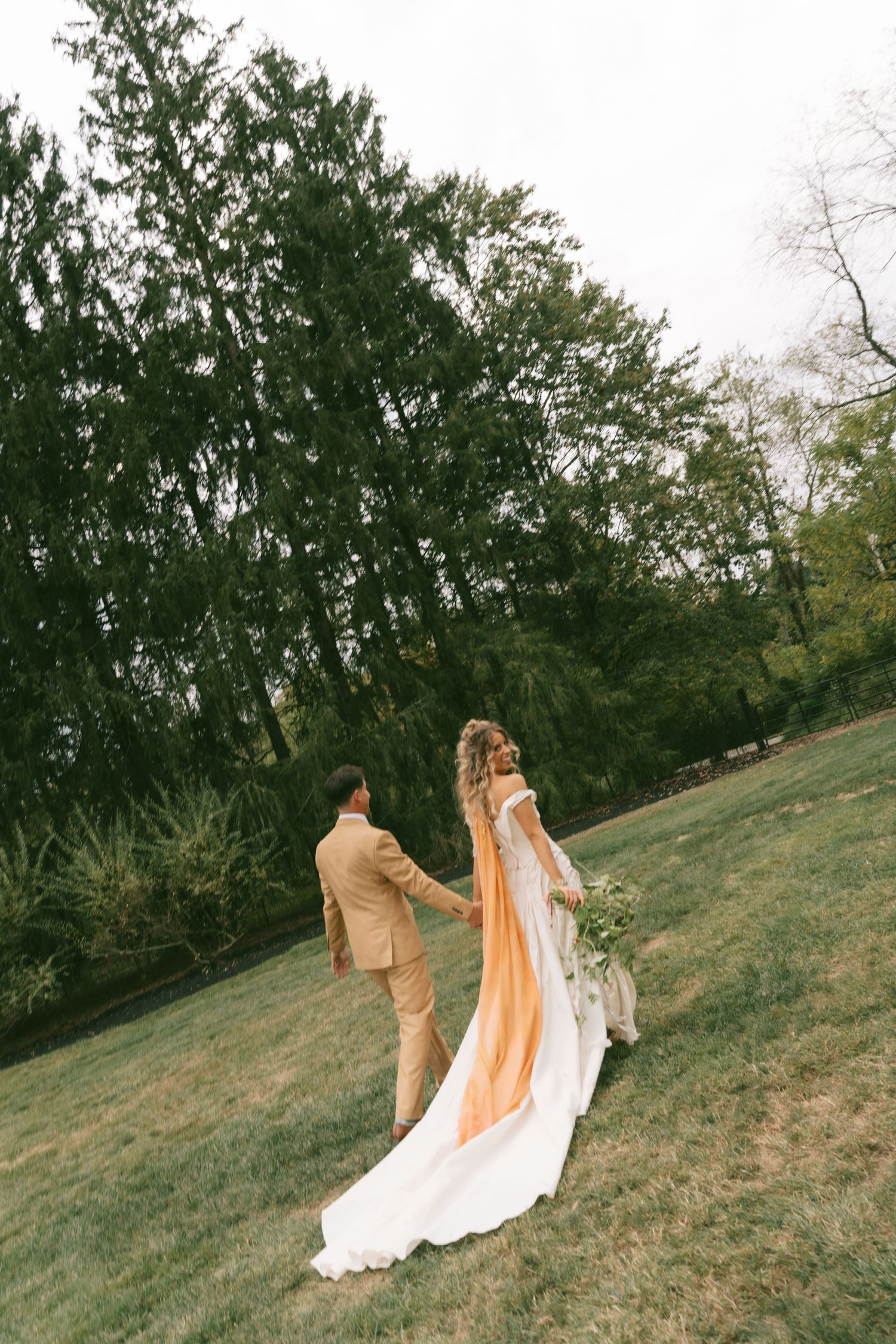 A couple in wedding attire walking together on a grassy hillside surrounded by trees.