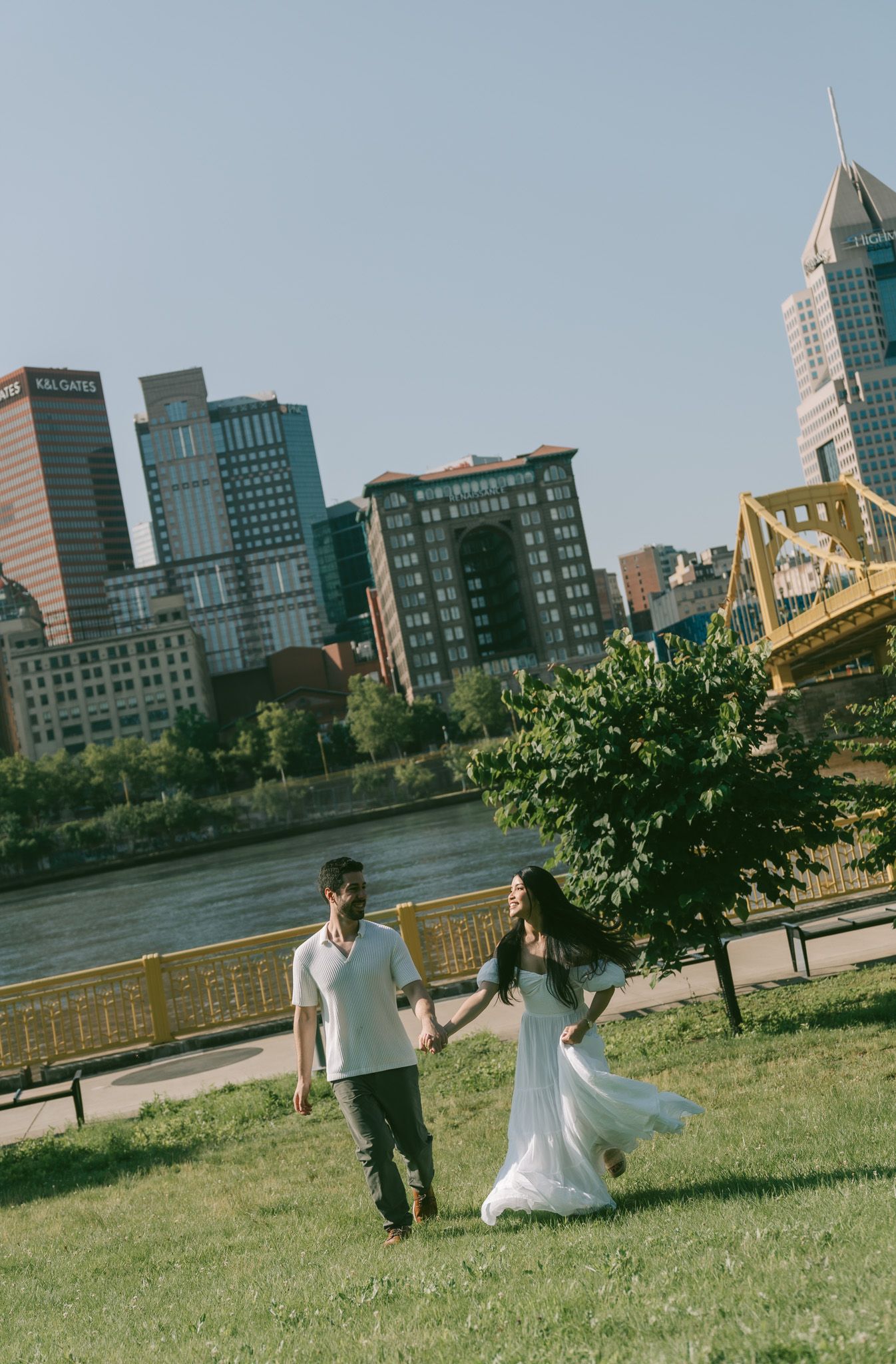 A couple walks hand in hand on green grass along the Pittsburgh riverfront with city skyscrapers in the background in north shore park