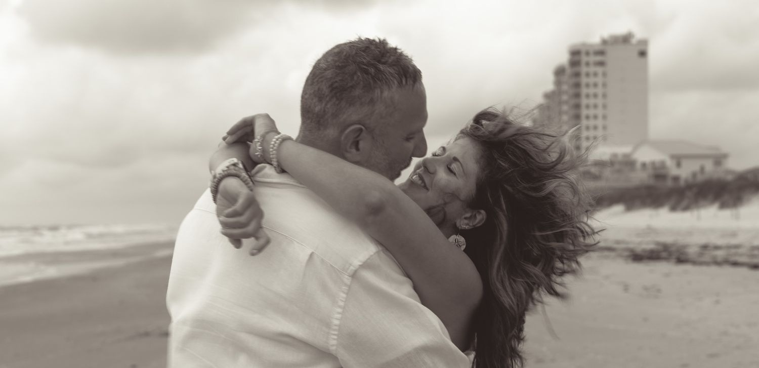 Romantic couple embracing on a cloudy beach with high-rise building in the background.