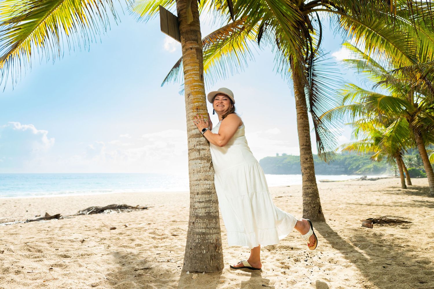 Woman in white dress and hat leans against palm tree on sunny tropical beach with blue sky.